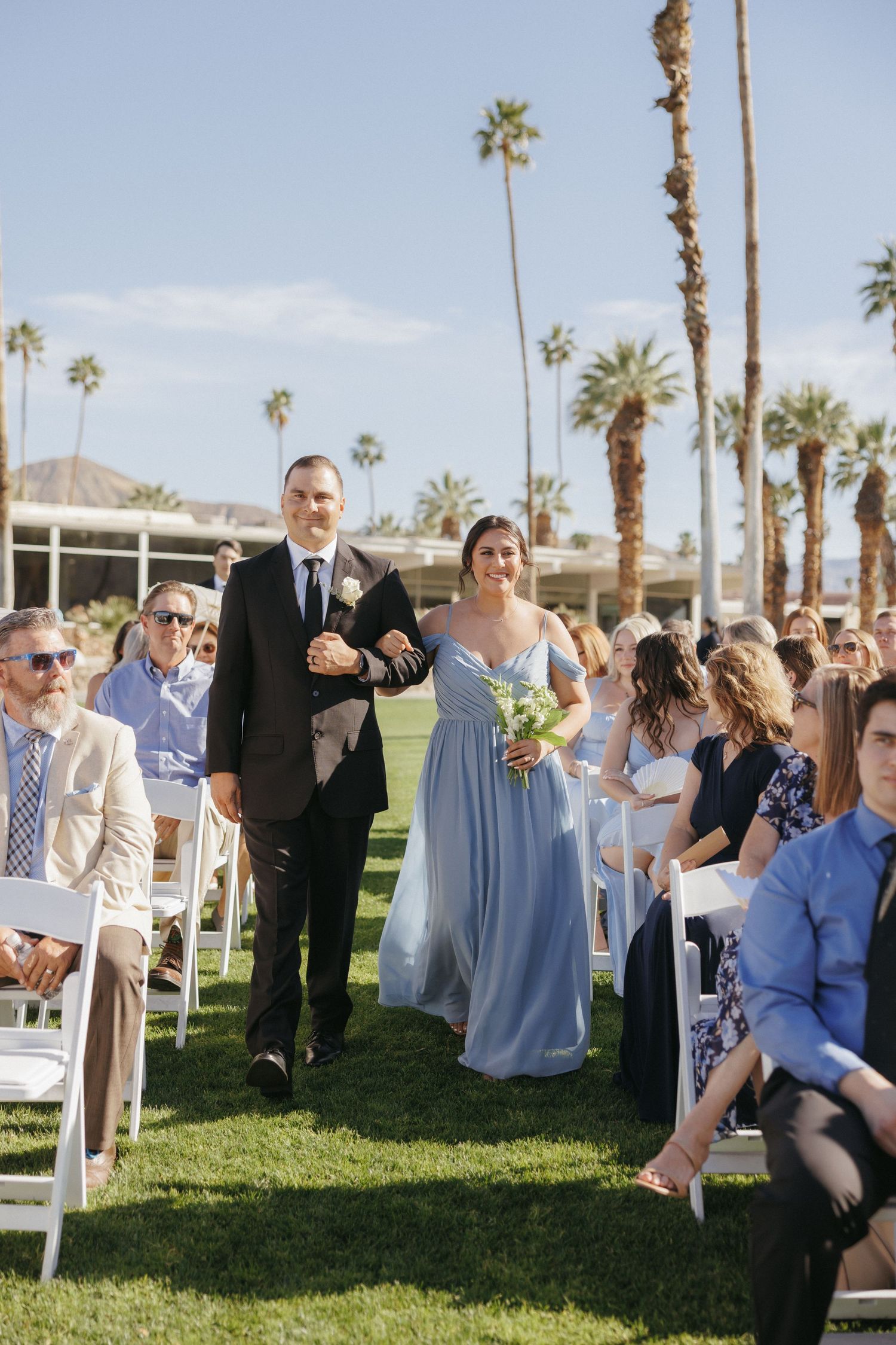 Bridesmaid in blue gown walks down outdoor aisle with groomsman at Palm Springs wedding.