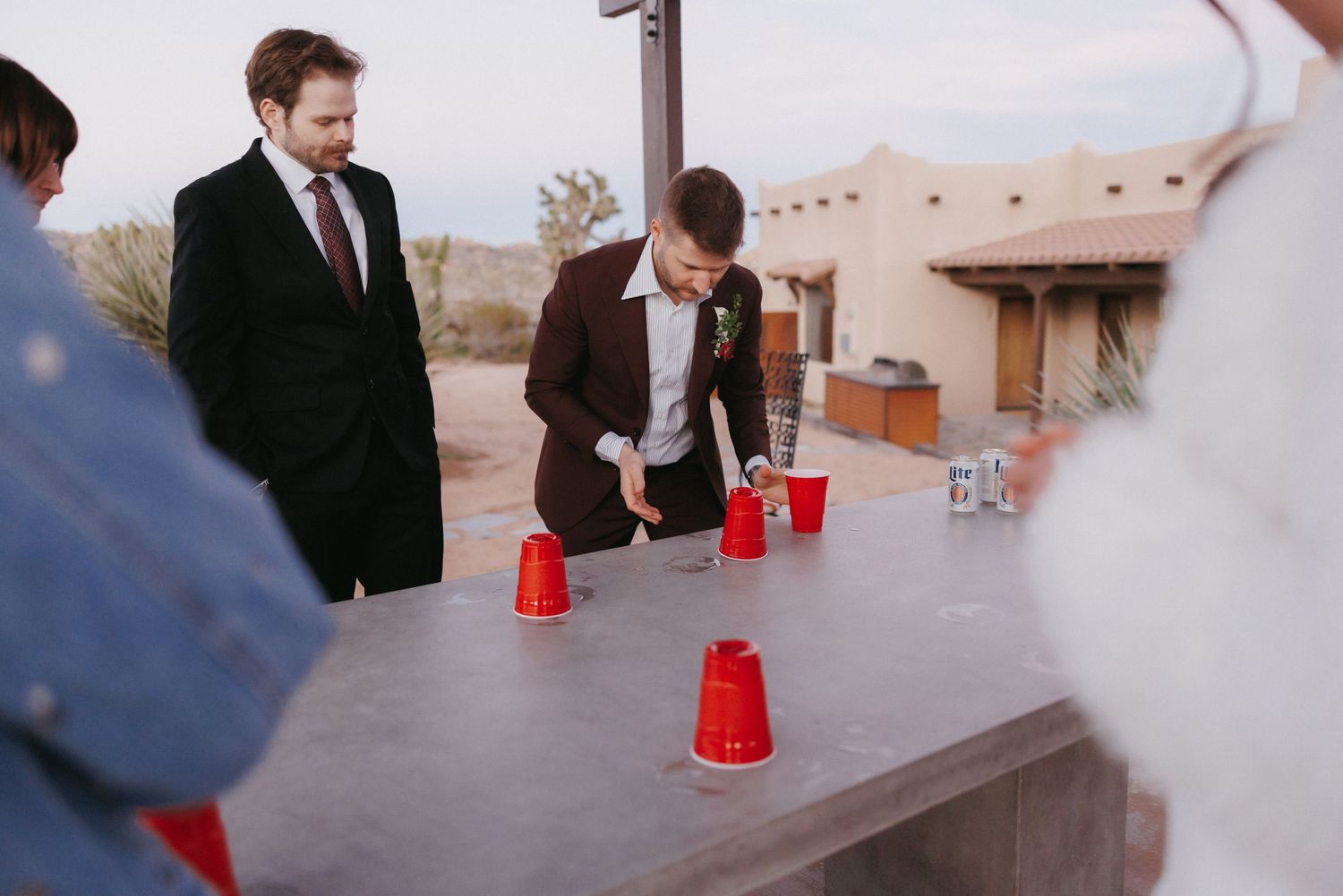 Groom in brown suit plays beer pong with red cups at outdoor wedding reception.