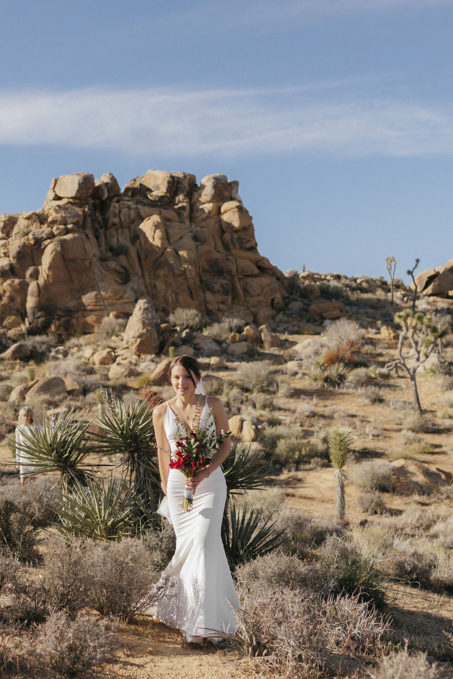 Bride in white gown holds bouquet while walking through Joshua Tree desert landscape.