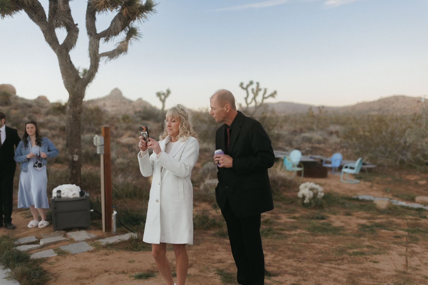 Two wedding guests share a toast outdoors at dusk with Joshua Tree desert landscape behind them.