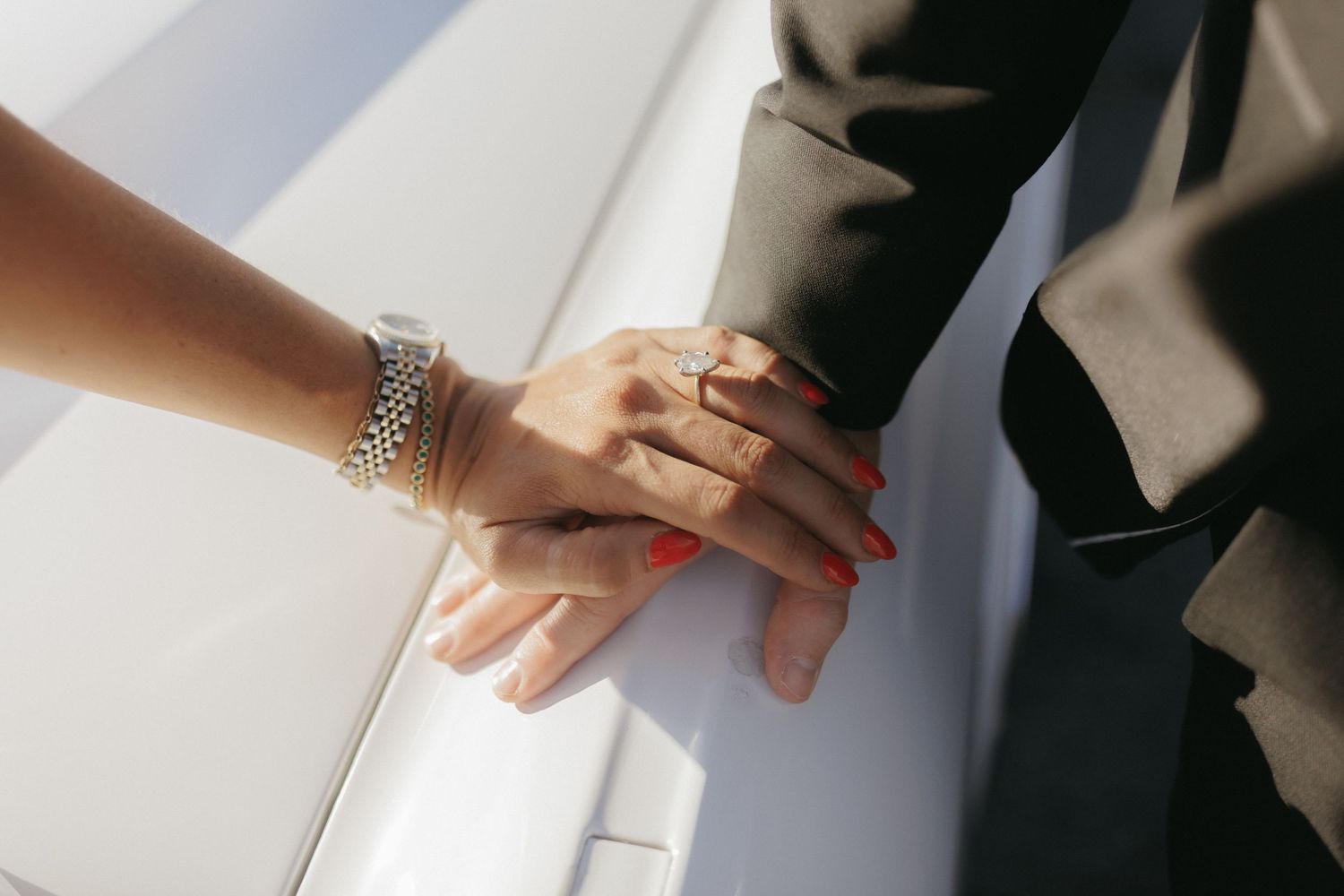 Close-up of bride and groom holding hands, bride wearing silver bracelet and red nail polish on white car.