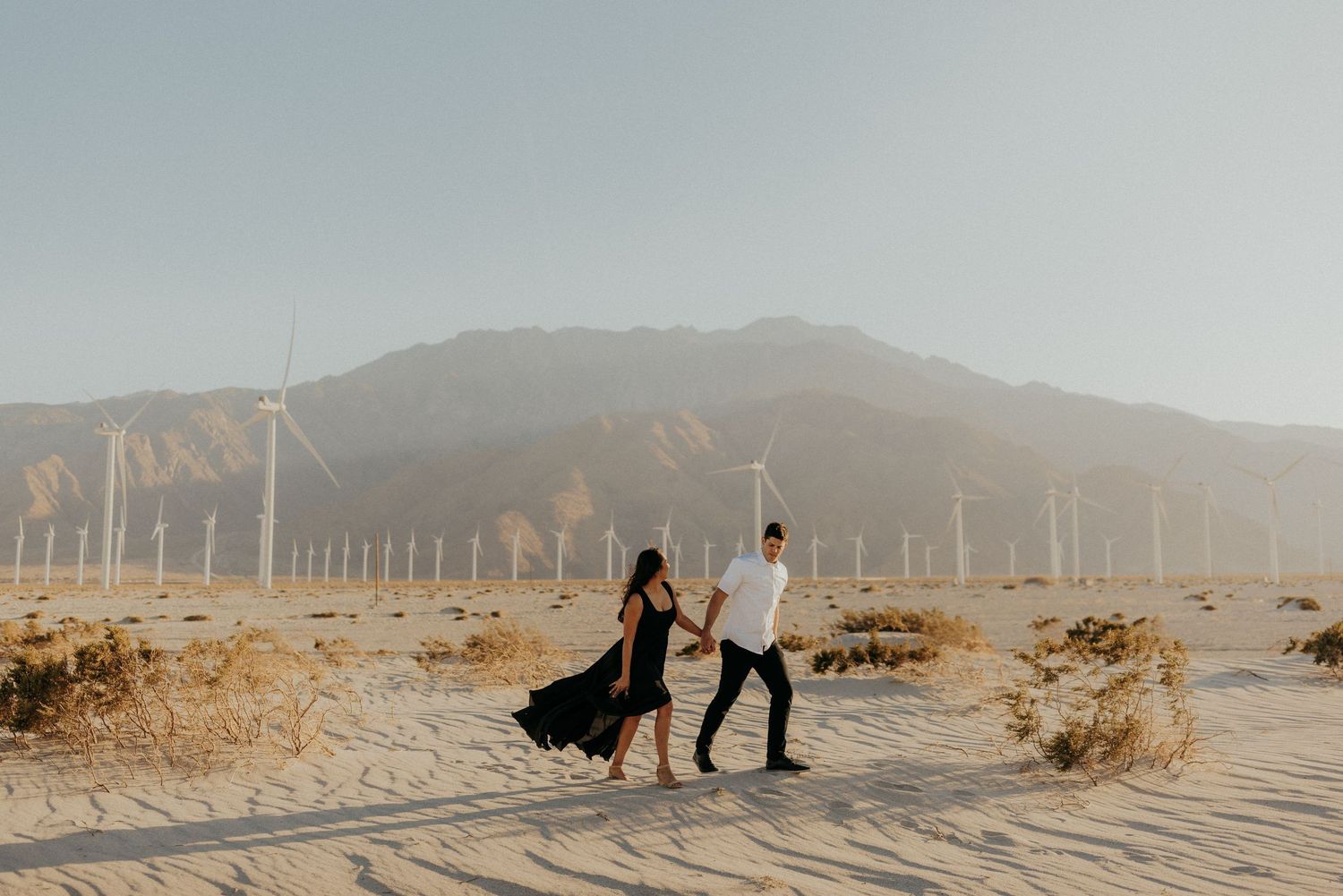 Panoramic misty desert landscape dotted with wind turbines stretches across hills beneath a hazy sky.