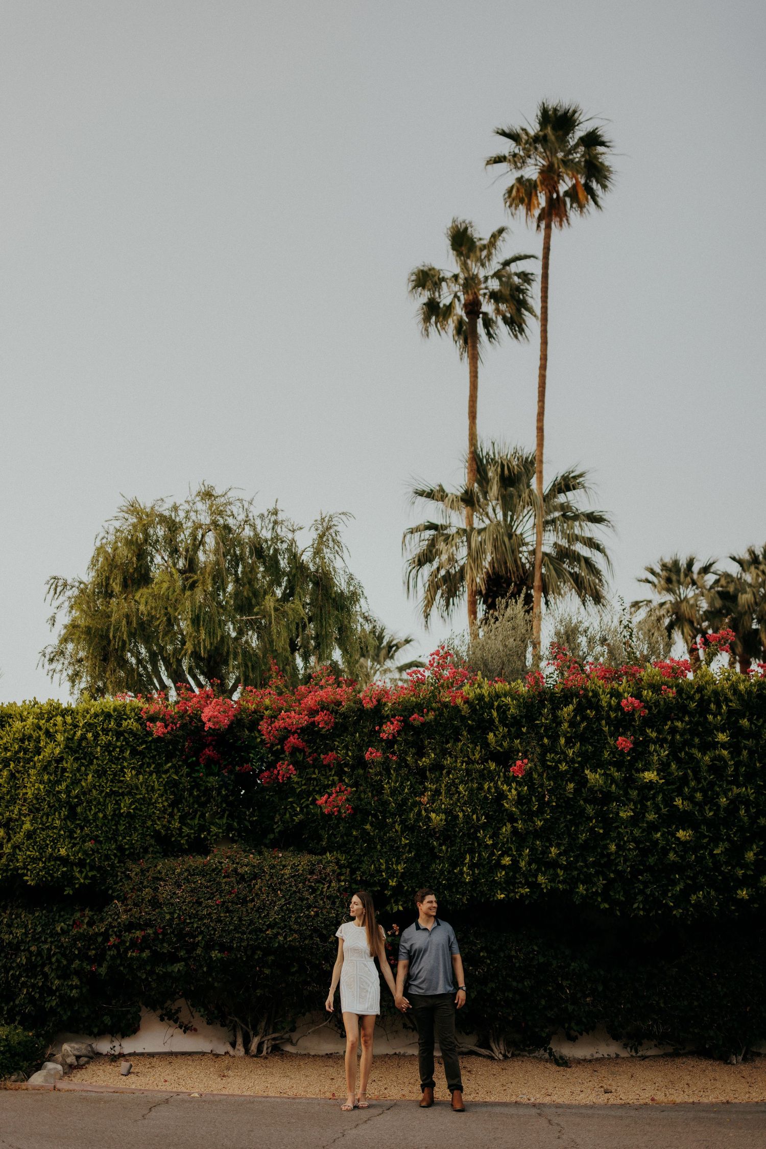 Couple walking in front of lush hedgerow with palm trees and pink bougainvillea flowers in background.