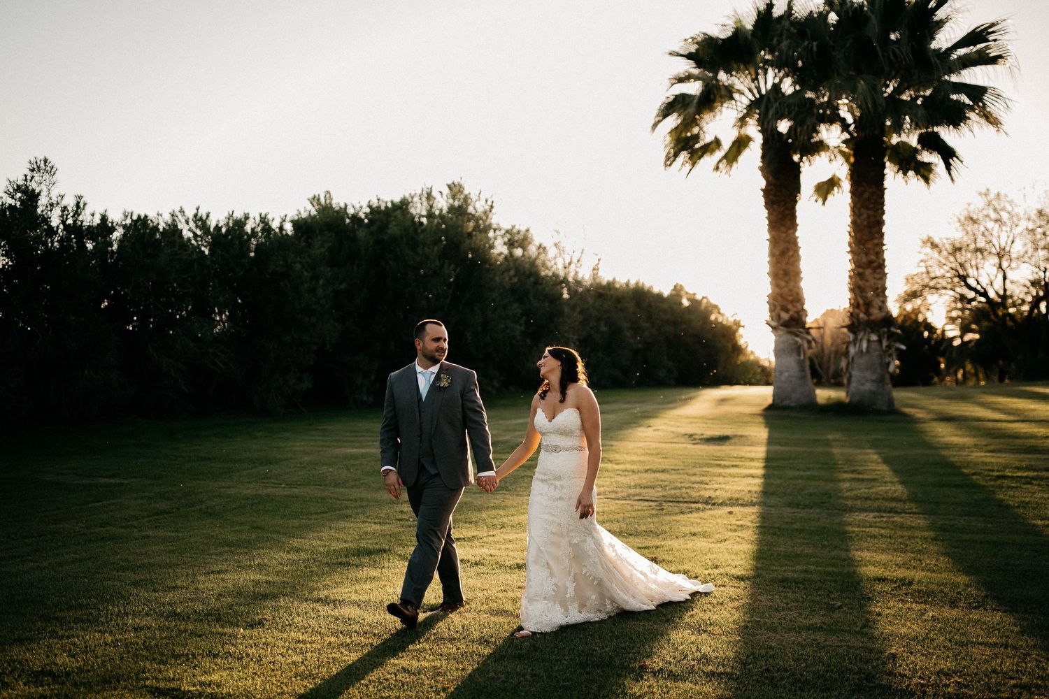 Wedding couple walking through golden sunlit grass on their special day at sunset creating romantic silhouettes.