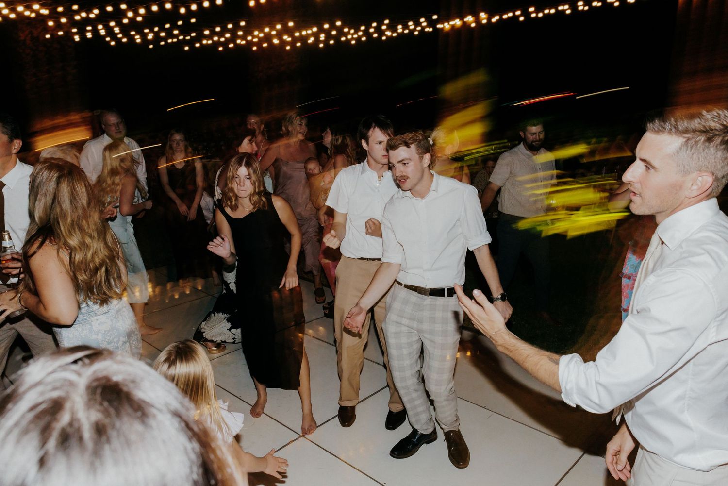 People dancing together on a dance floor under glowing string lights at an evening celebration party.