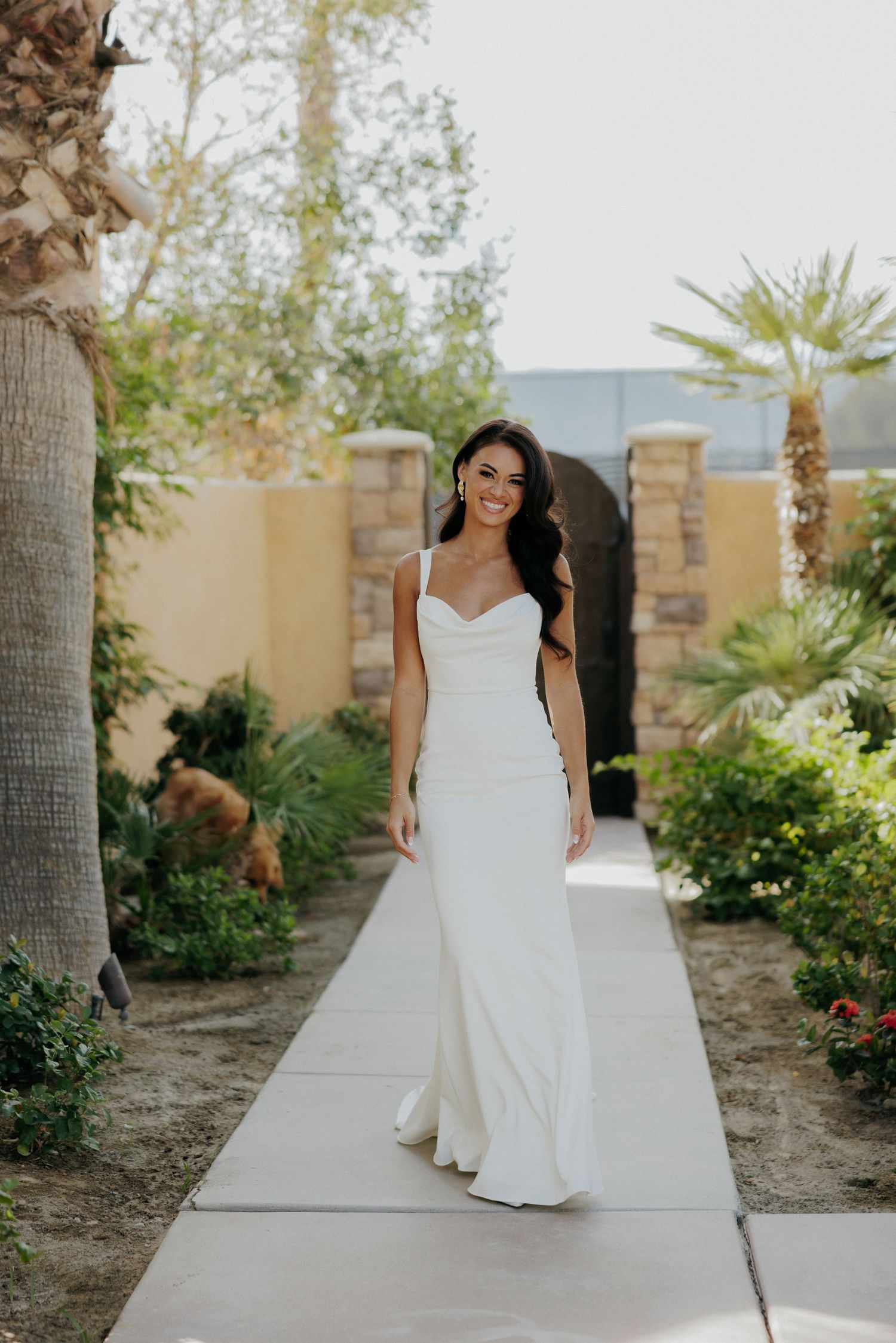 Bride in flowing white wedding dress sits gracefully on concrete ledge with palm trees and desert landscaping in background.