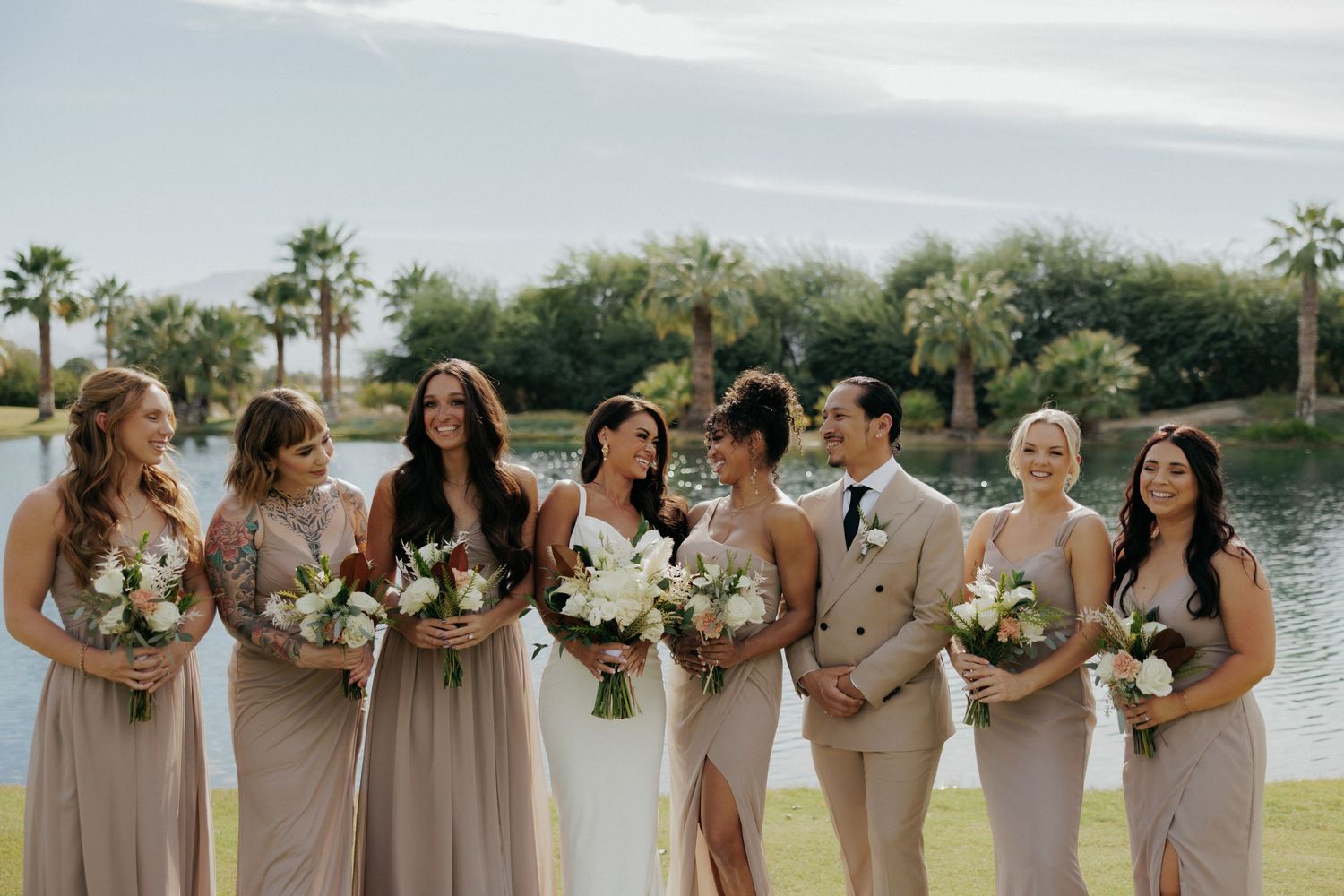 Row of bridesmaids in neutral-toned dresses stand with bouquets against waterfront backdrop.