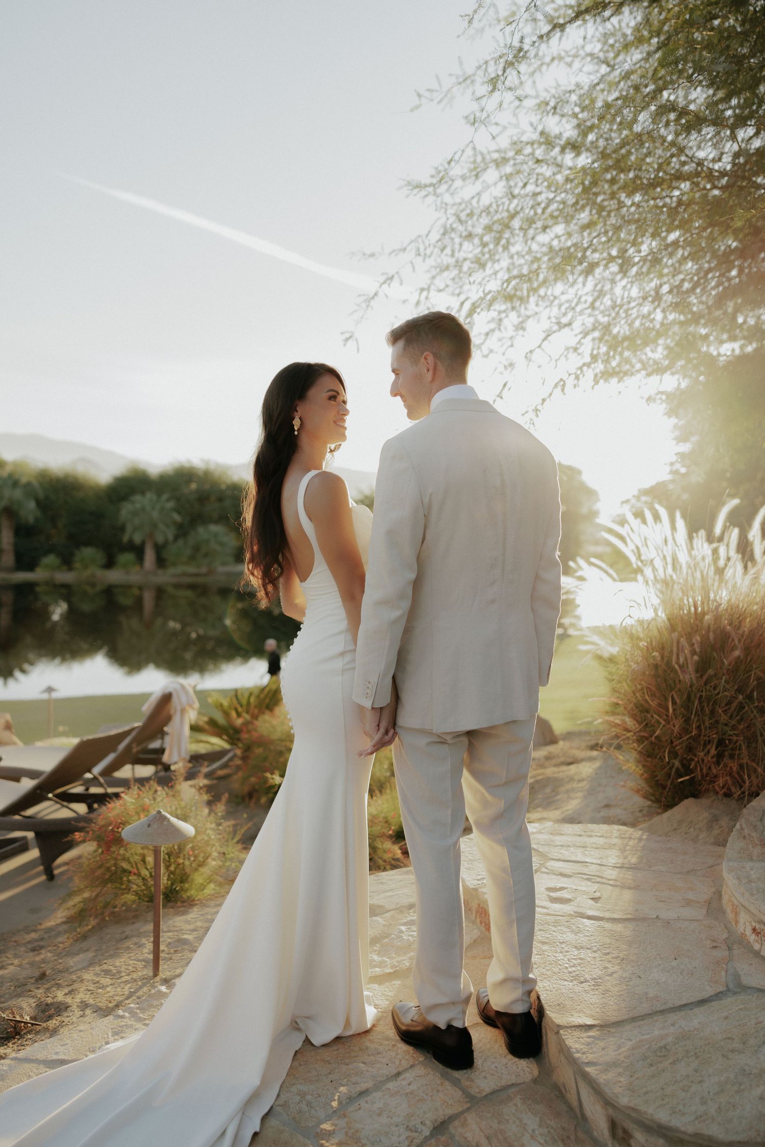 A couple in wedding attire embraces by a tranquil waterside path during golden hour sunset.