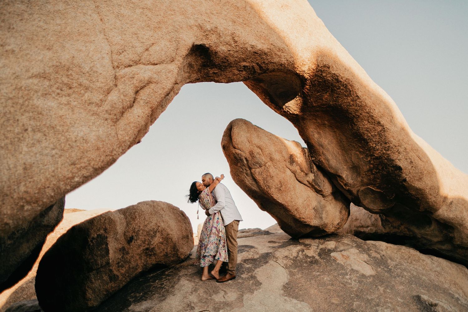 Couple embraces beneath massive desert rock formations at Joshua Tree National Park during sunset.