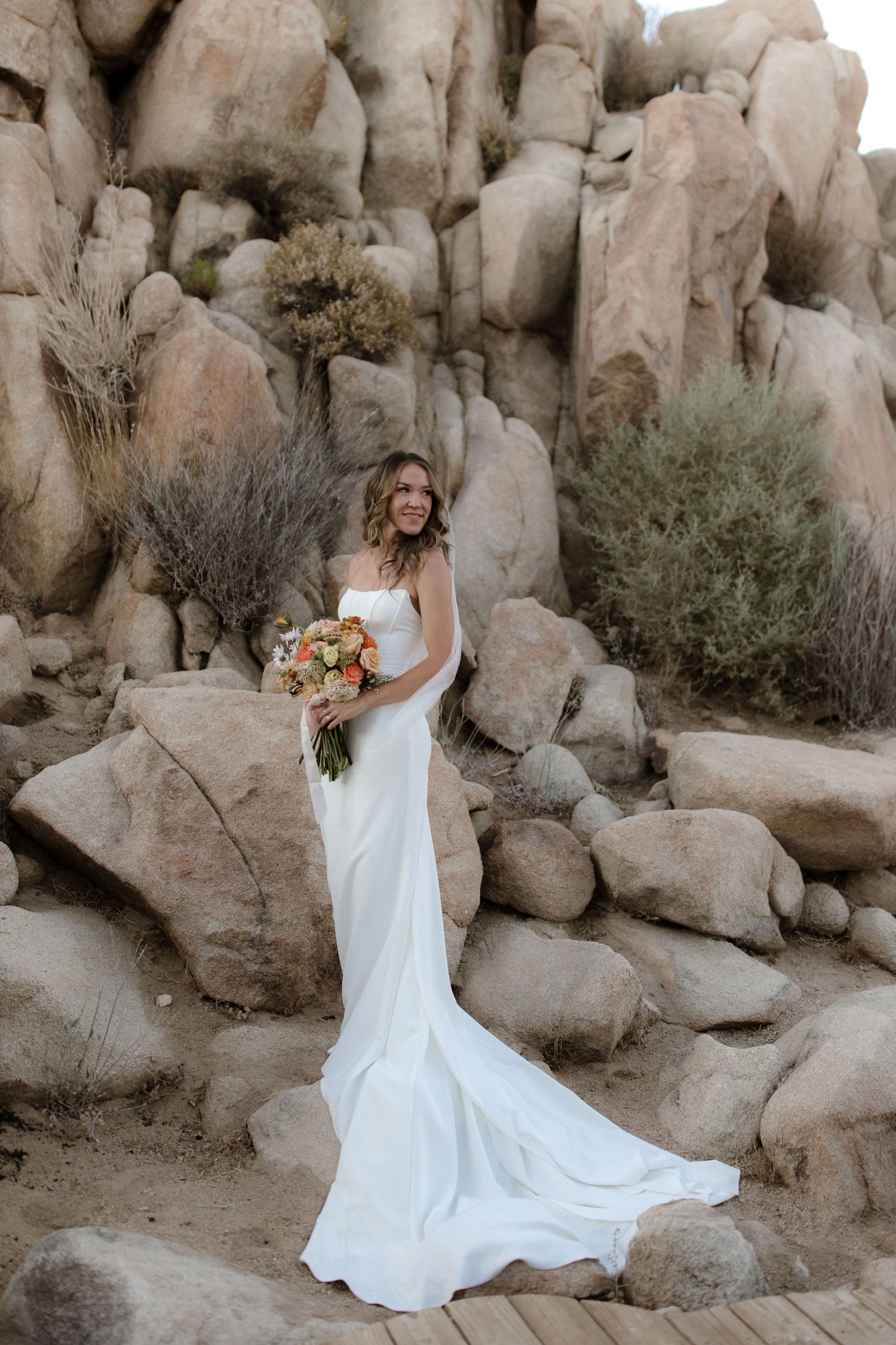 A bride in a flowing white gown poses elegantly against desert rock formations at sunset.