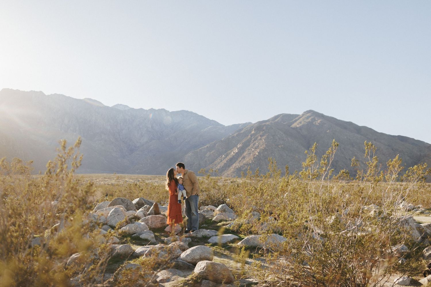 Couple embracing in a sunlit desert landscape with rocky terrain and mountain backdrop under a clear blue sky.
