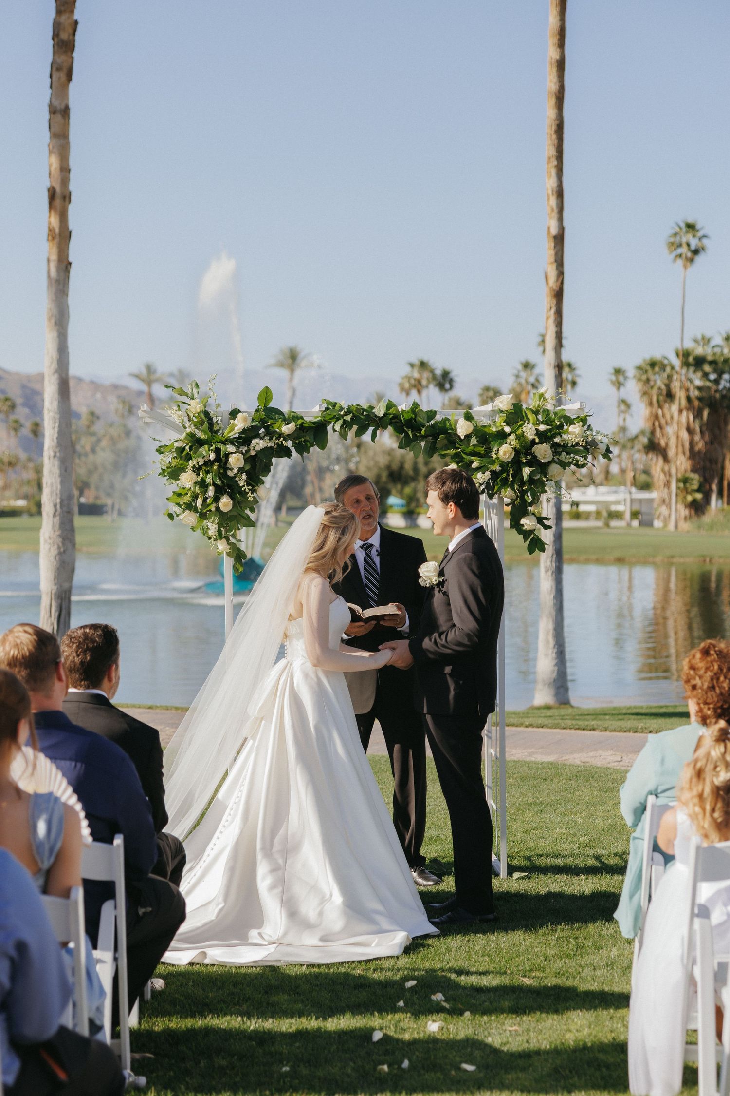 Bride and groom exchanging vows at outdoor lakeside ceremony with palm trees and floral arch.