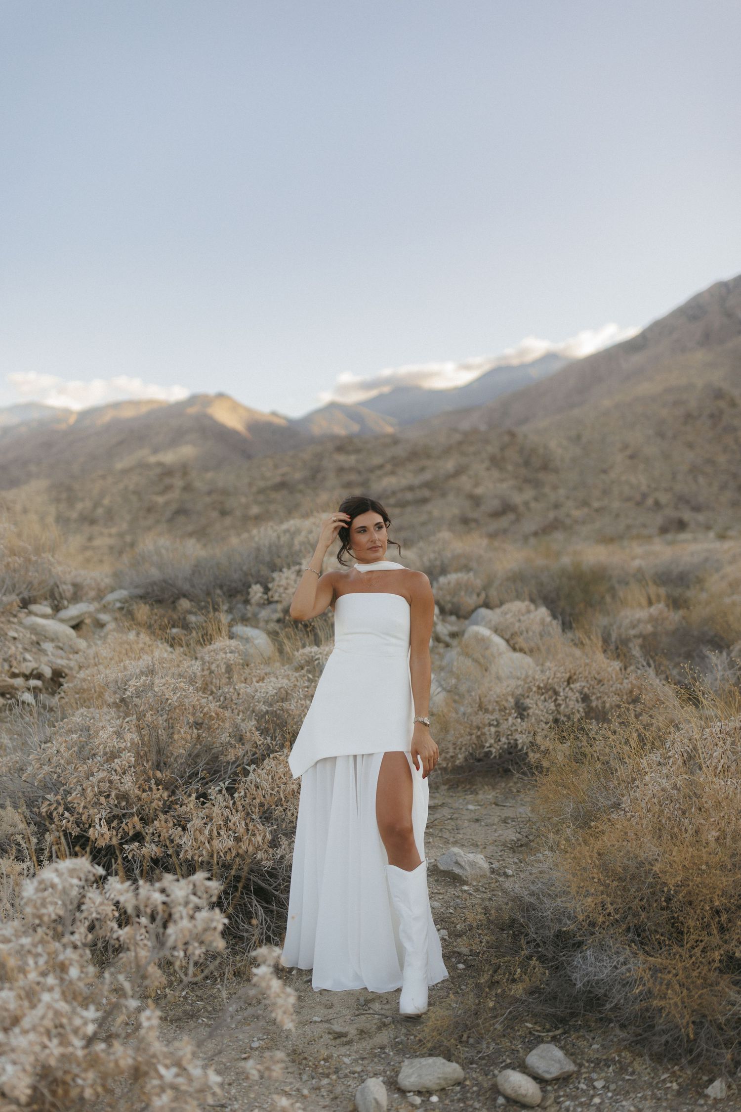 Bride in strapless white gown with high slit poses in desert landscape with rocky mountains at golden hour.