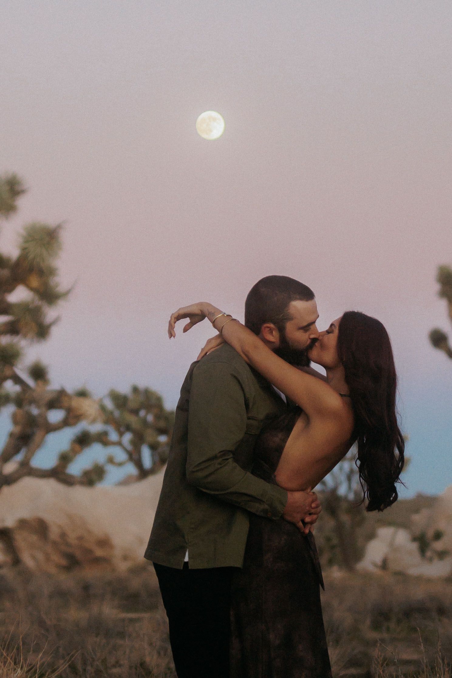 Couple kissing under a full moon at dusk in Joshua Tree desert landscape with Joshua trees and boulders.