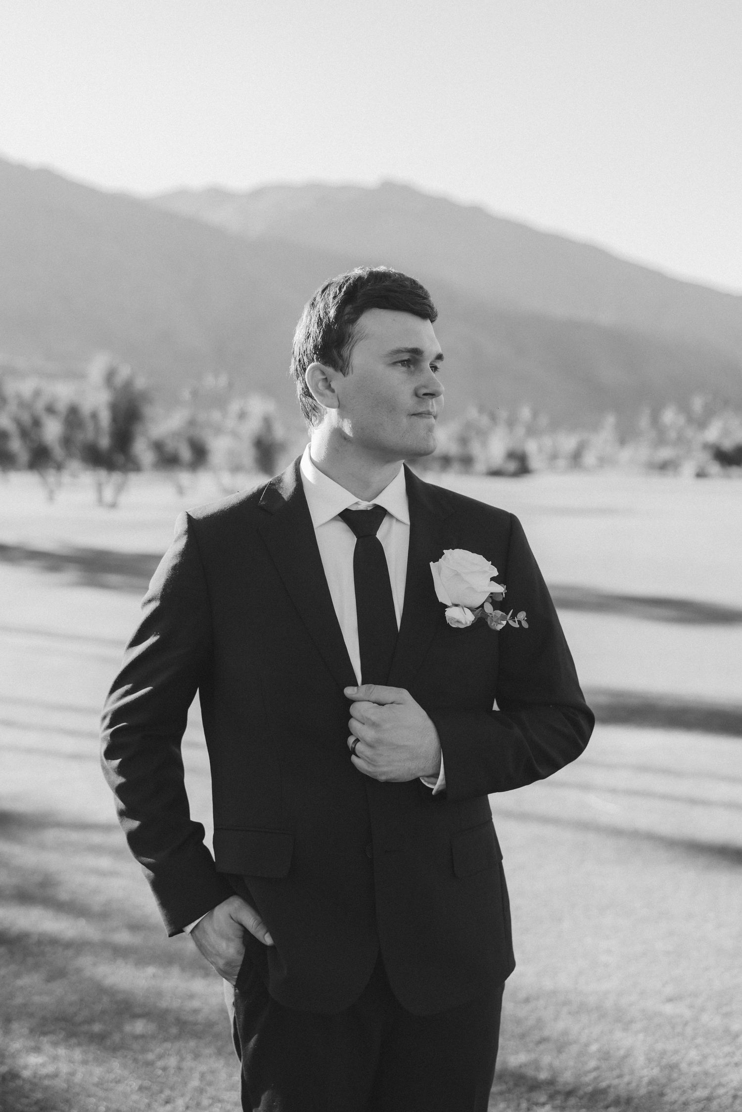 Handsome groom in black suit with white boutonniere poses outdoors in black and white photo.