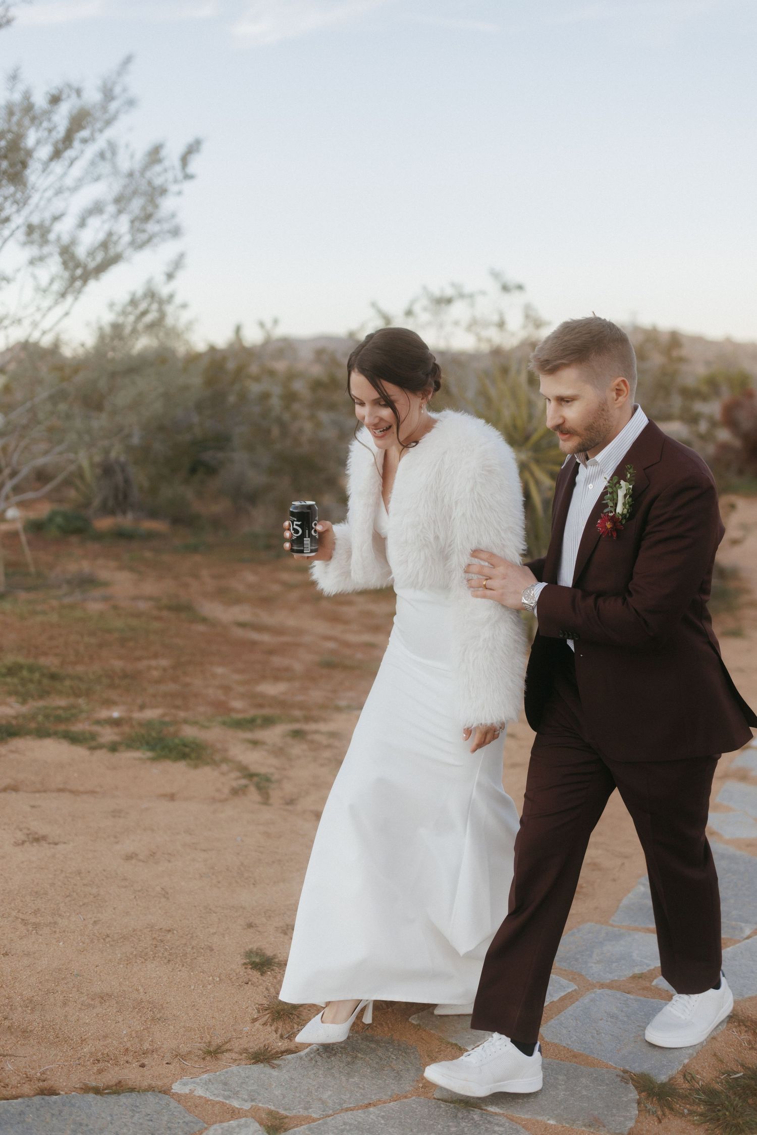 Bride in white fur wrap and groom in burgundy suit walk together at desert wedding.