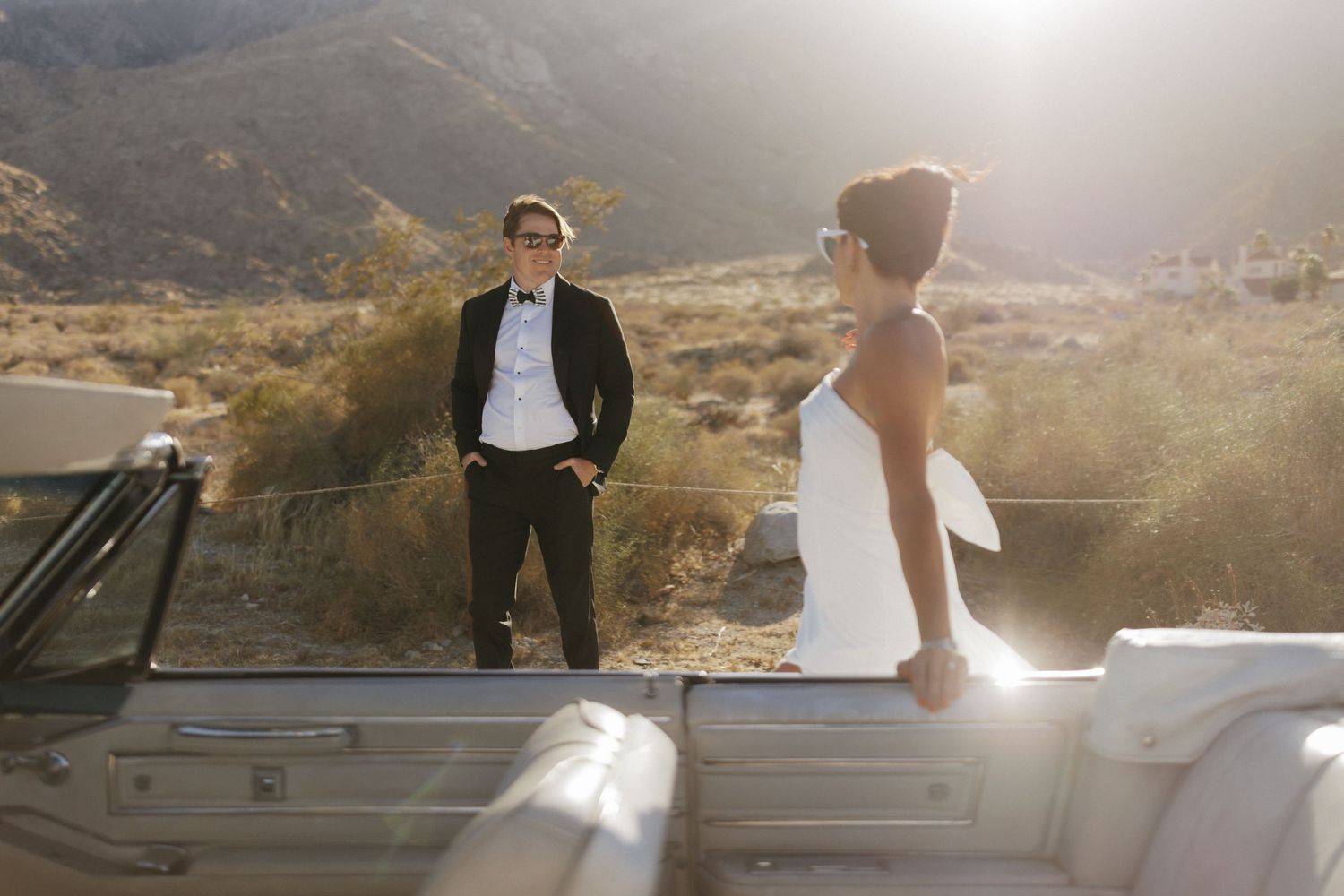 Bride and groom in wedding attire stand beside a white convertible with sunlit desert mountains backdrop.