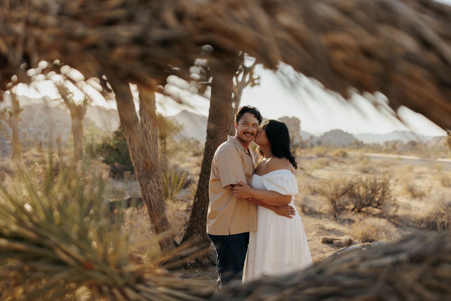 Couple embracing under a Joshua tree in a desert landscape, woman in white dress, man in beige shirt.