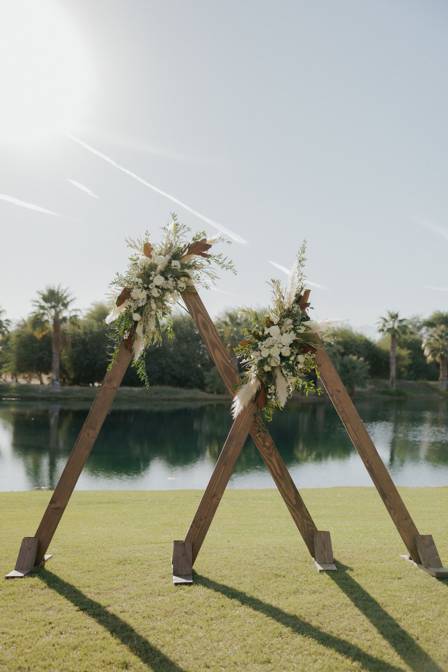 Wooden triangular wedding arch decorated with floral arrangements creates dramatic shadows on a lakeside lawn.