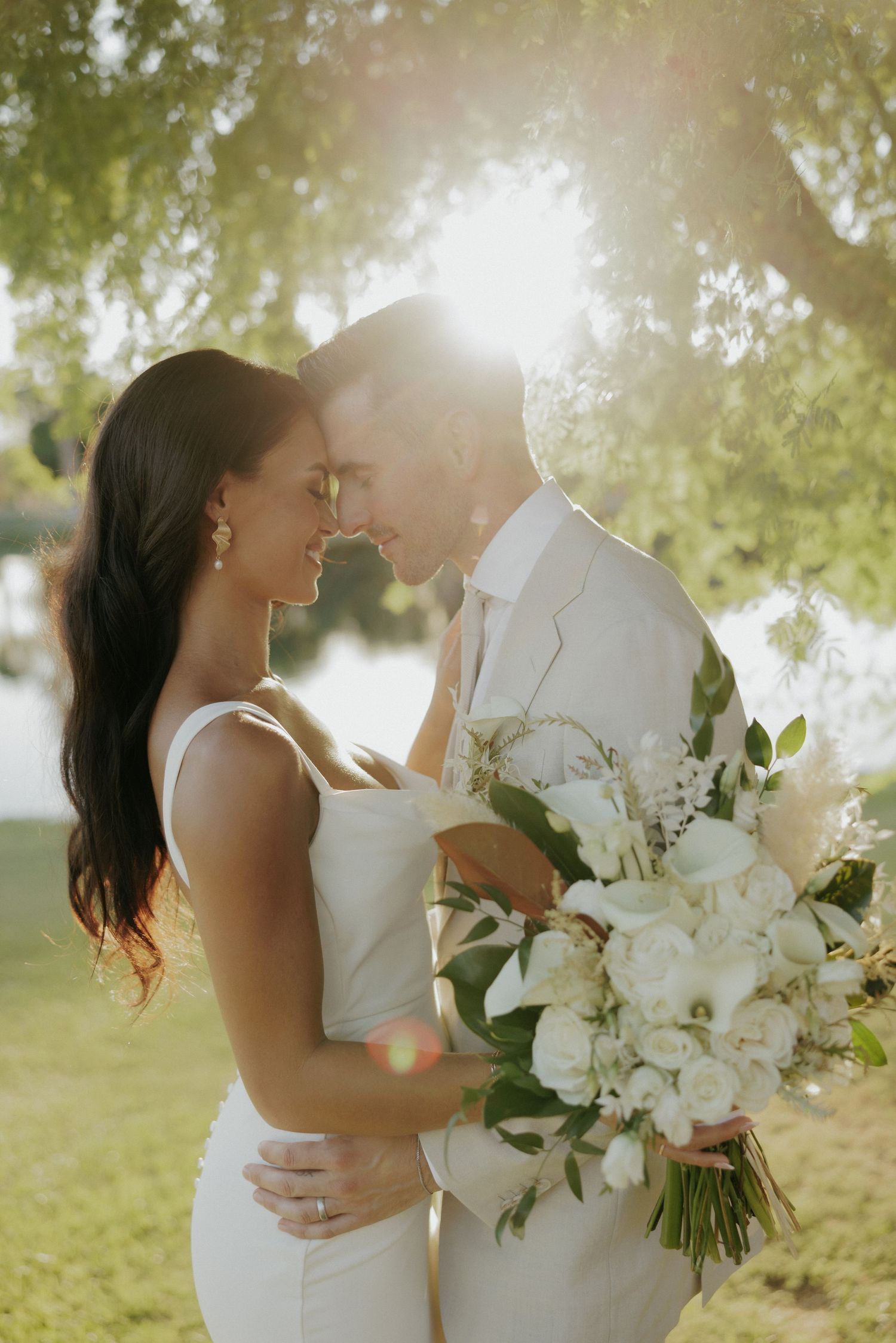 A romantic outdoor wedding couple embraces in soft sunlight while holding a bouquet of white flowers.