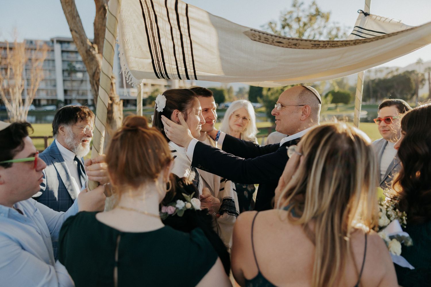 Jewish wedding ceremony under chuppah with guests gathering around the couple in an outdoor setting.