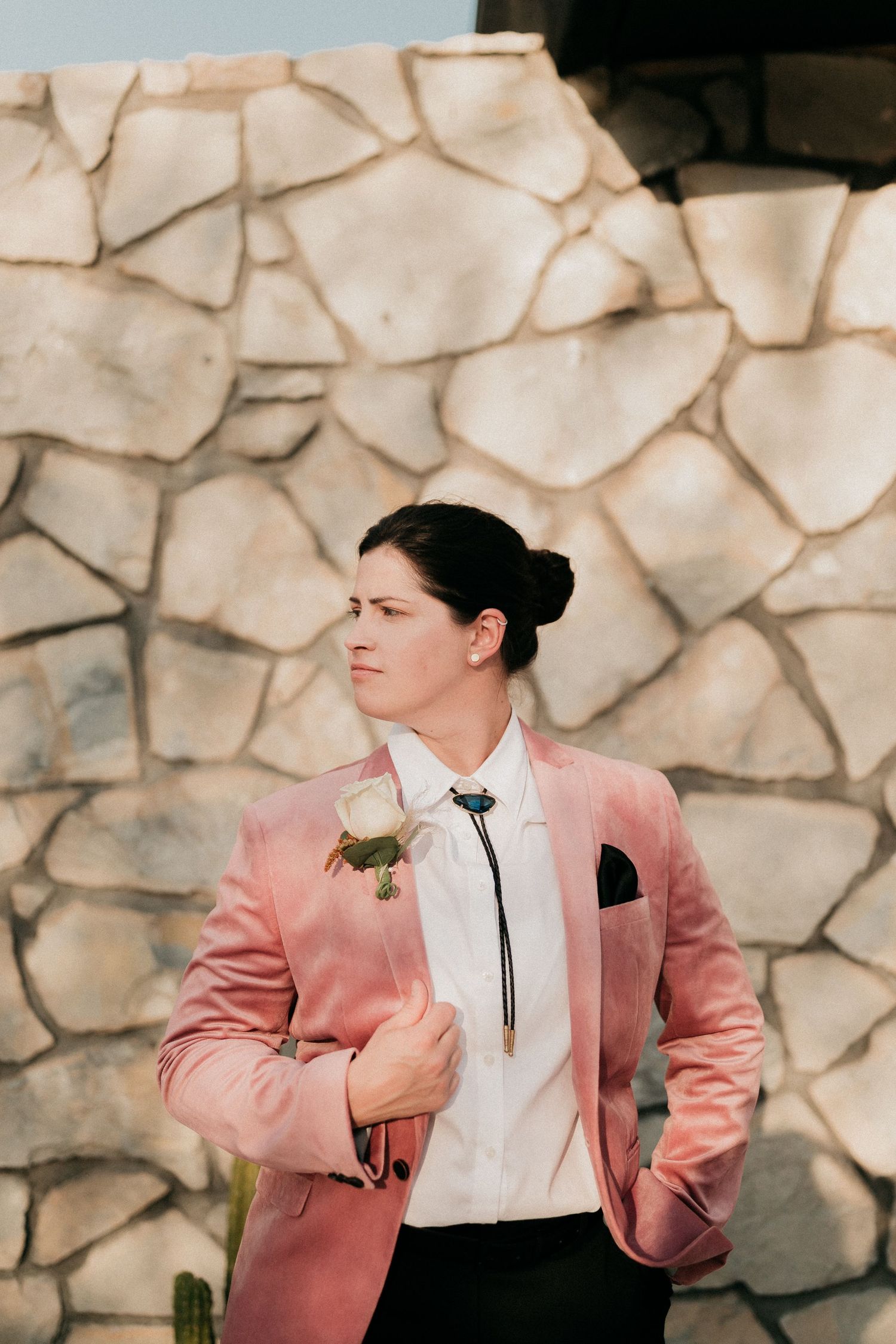 Person wearing a pink suit jacket and white shirt sits against a stone wall background.