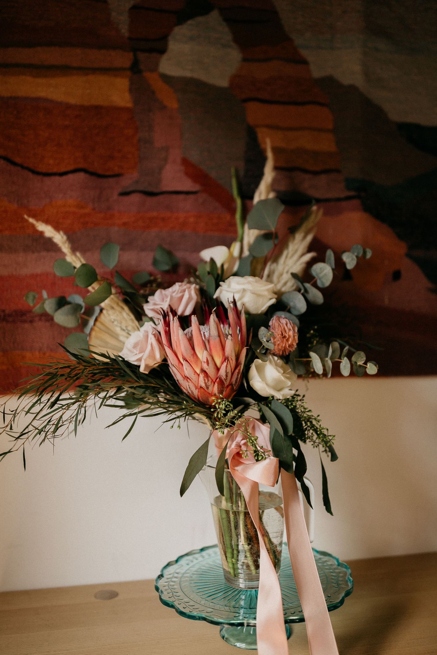 Delicate floral arrangement featuring pink protea and eucalyptus on a turquoise glass pedestal against a wooden surface.
