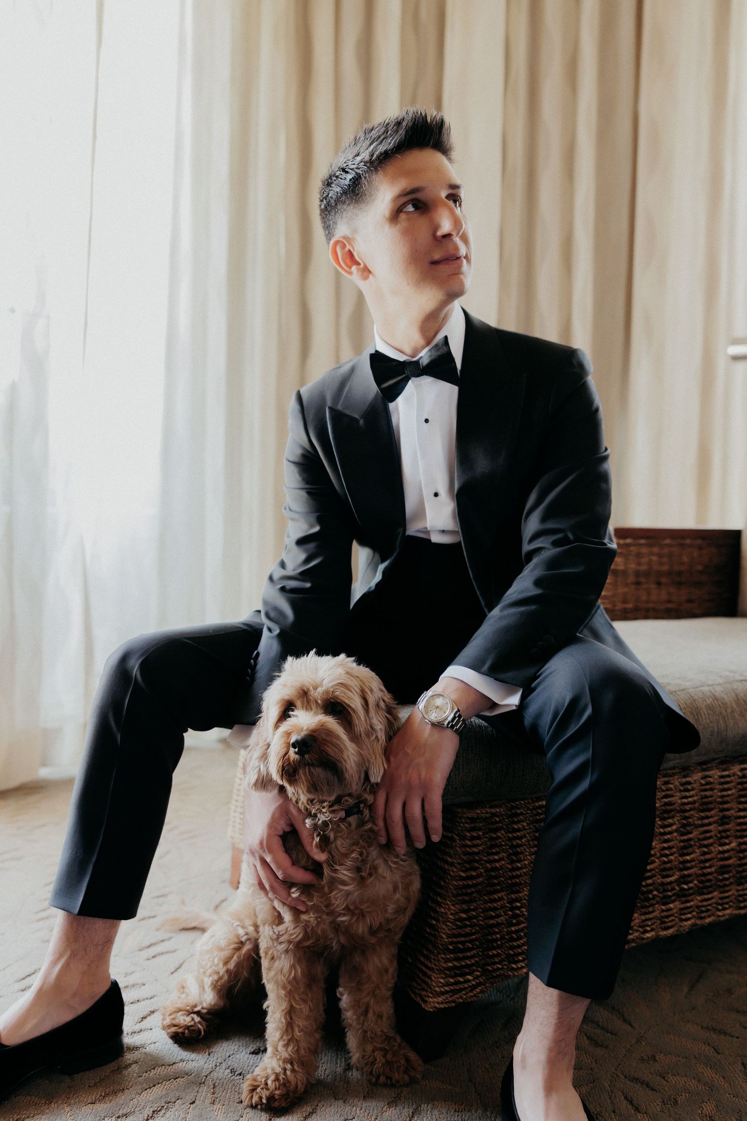 A person in a black tuxedo sits on rattan furniture with a small brown dog.