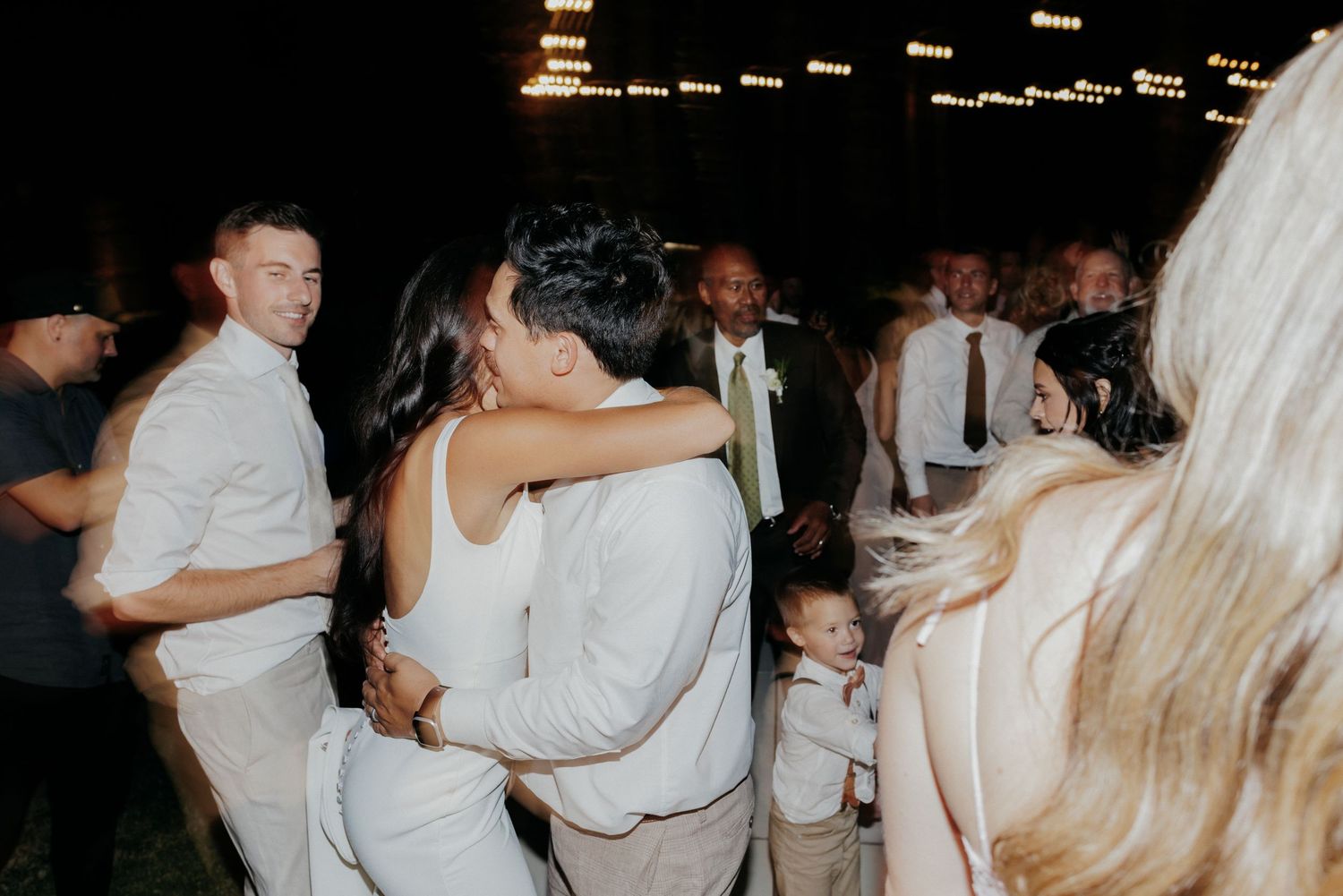 Wedding party enjoying dance floor moments with string lights glowing overhead during evening celebration.