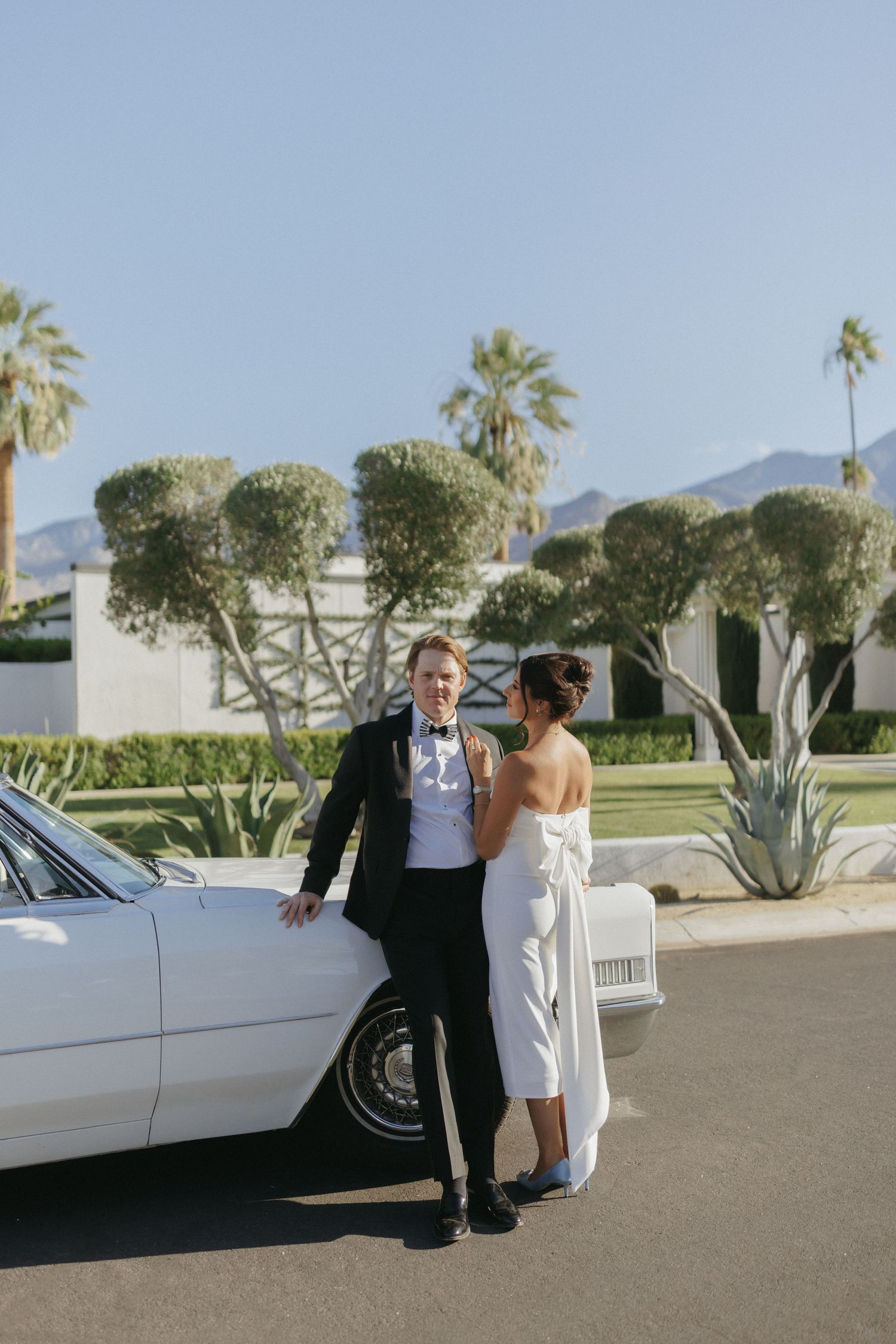 A bride and groom pose beside a classic white convertible with palm trees and mountains in Palm Springs.