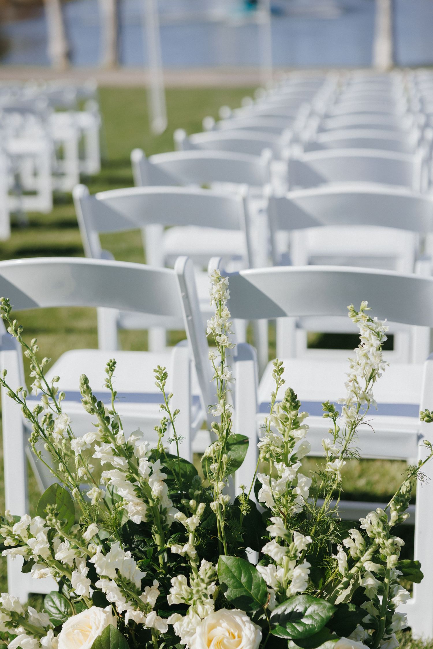 White folding chairs lined up outdoors with elegant white floral arrangements for a wedding ceremony.