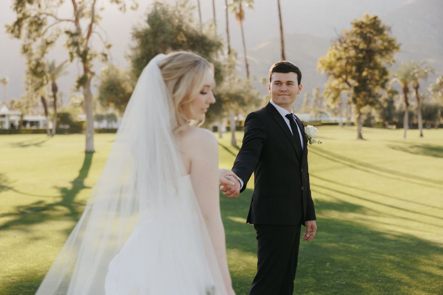 Bride and groom holding hands while walking on a green golf course with mountain views.