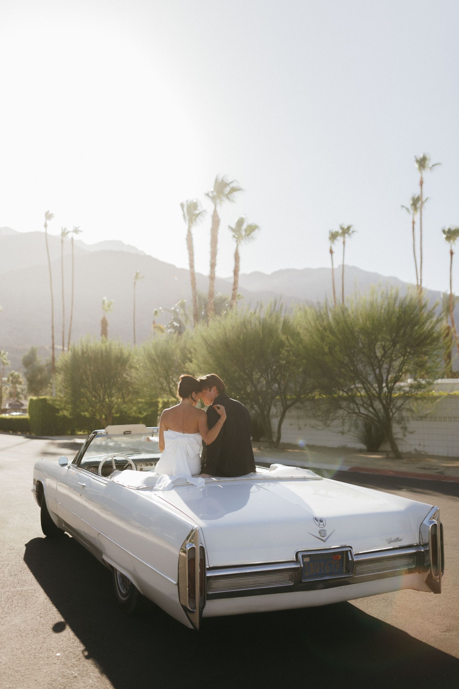Bride and groom kissing on a white vintage Cadillac convertible with palm trees and mountains in Palm Springs.