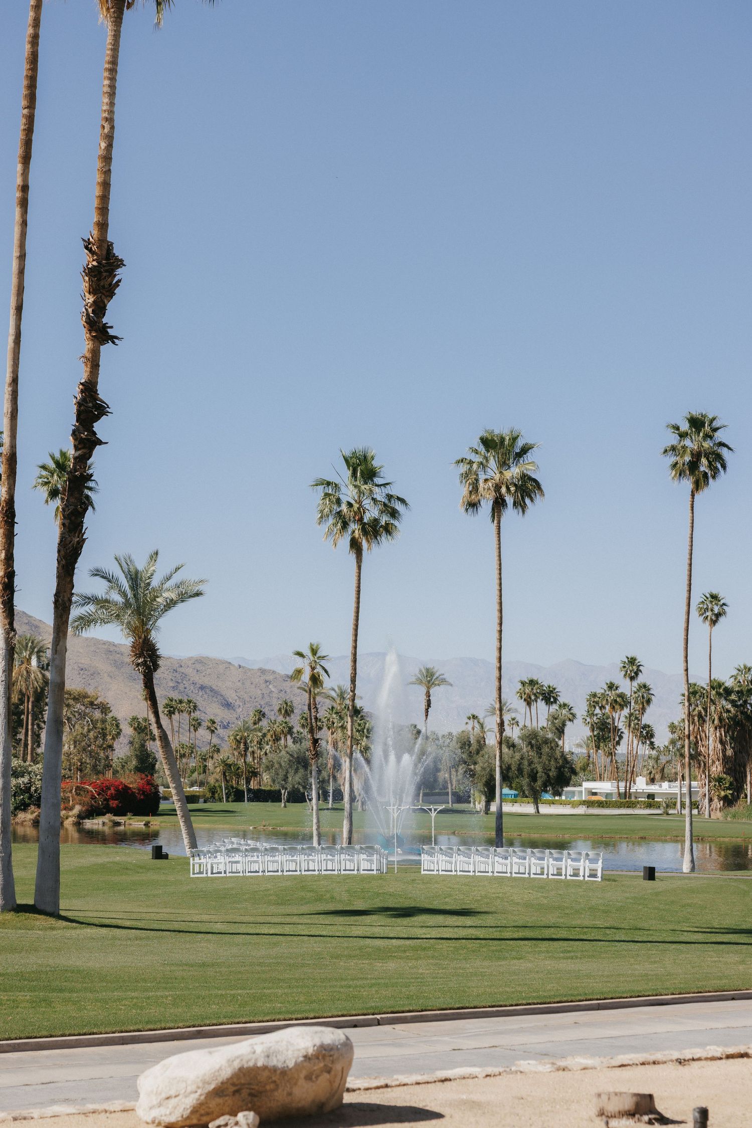 Outdoor wedding ceremony setup with white chairs near a fountain, surrounded by tall palm trees and mountains in Palm Springs.