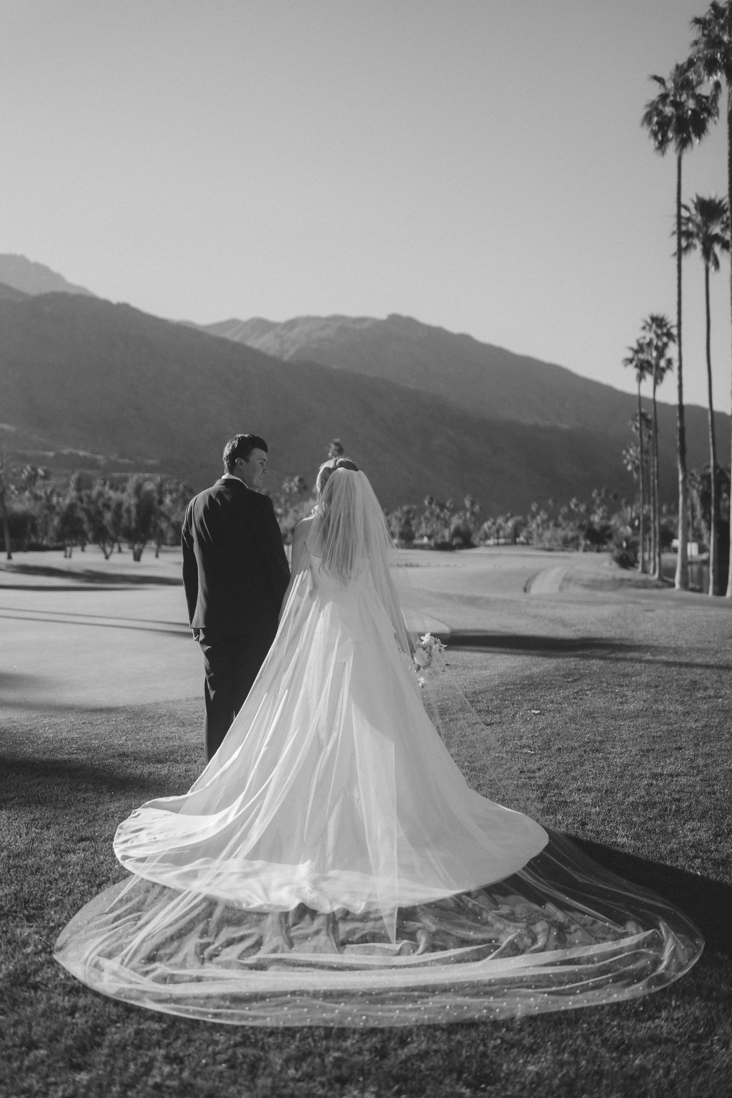 Black and white photo of bride and groom standing together with mountain and palm tree backdrop.
