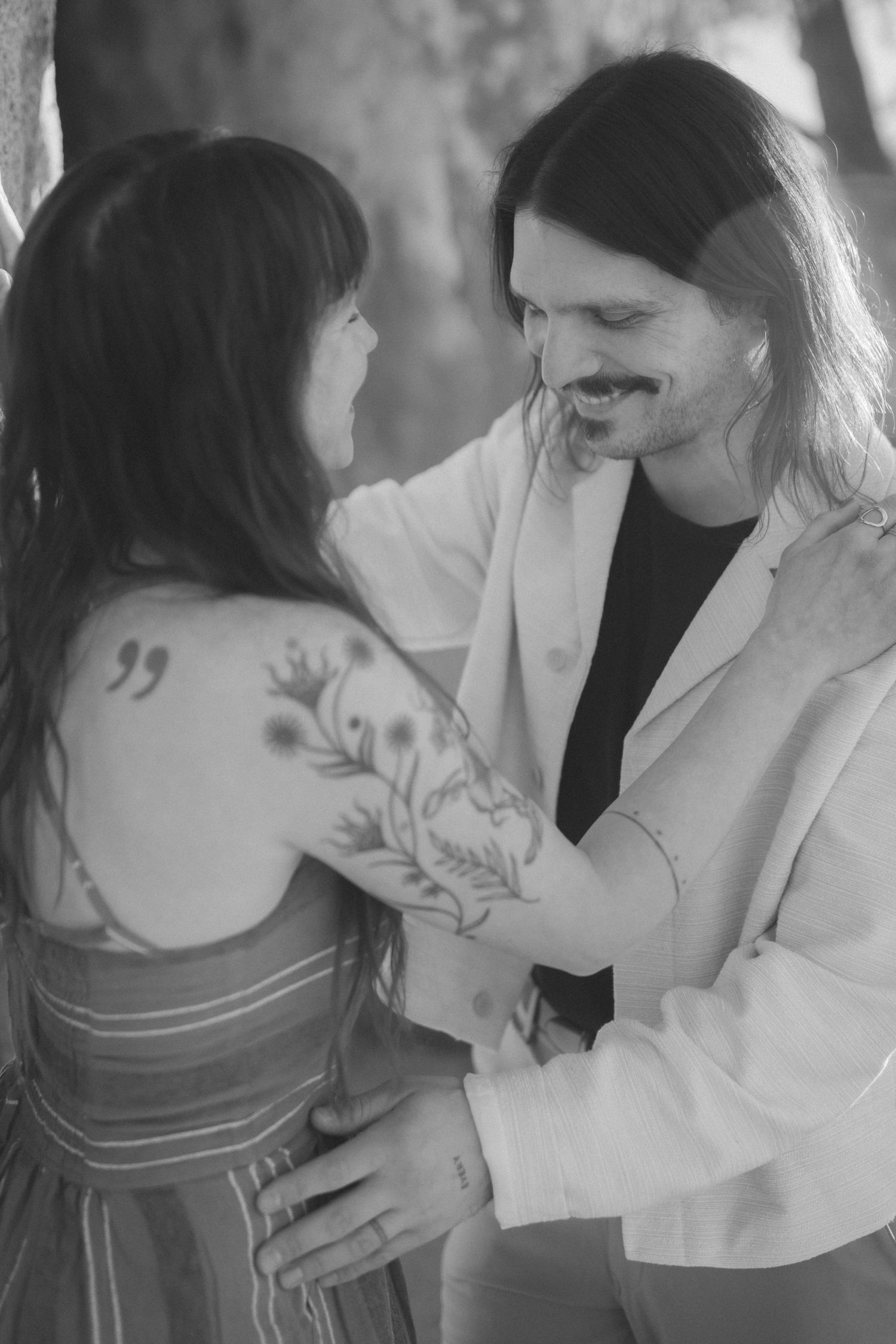 Black and white photo of a couple sharing an intimate moment, woman with floral tattoo on shoulder in striped dress.