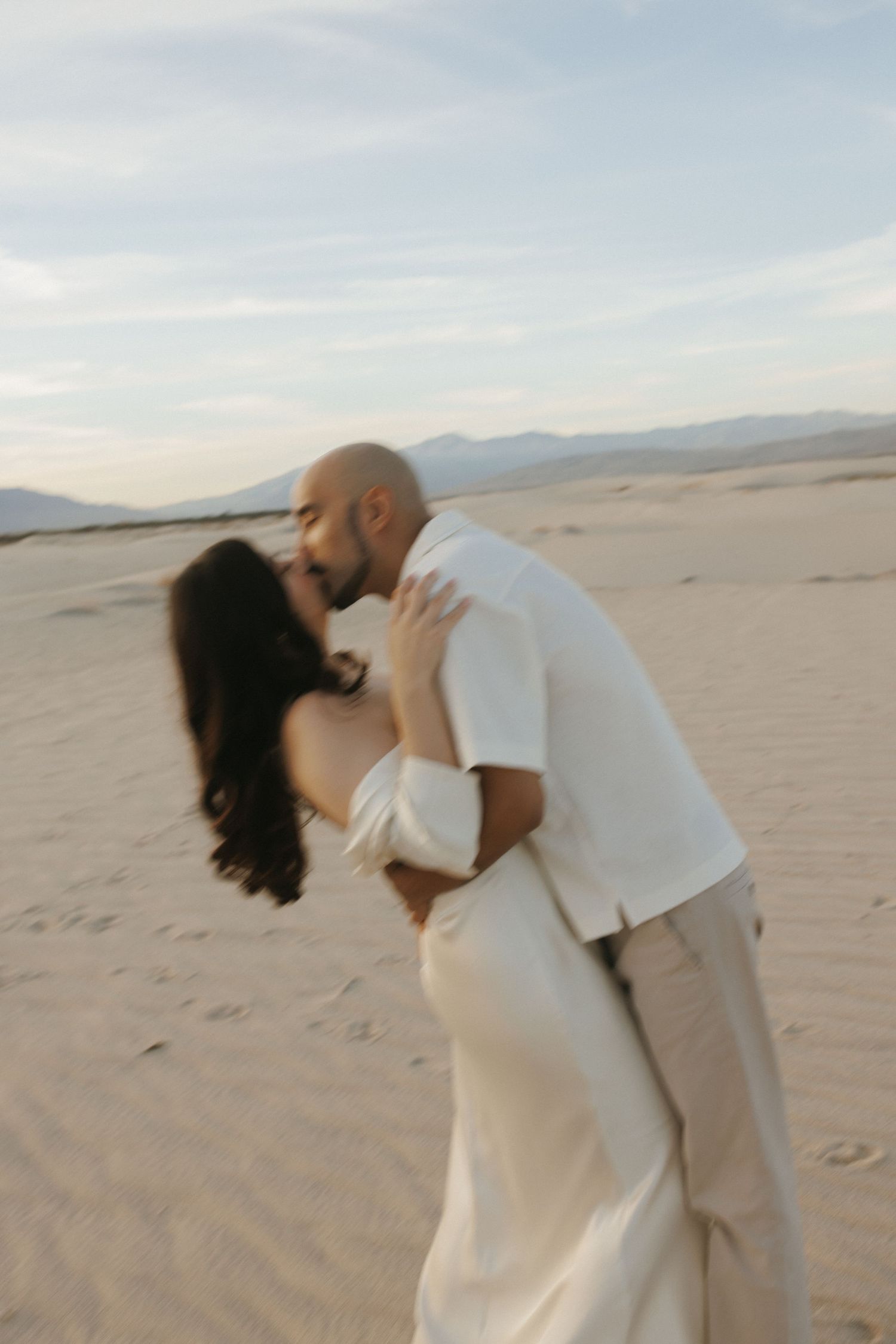 A couple shares a romantic kiss while embracing on sandy desert dunes at dusk, dressed in elegant cream and white attire.