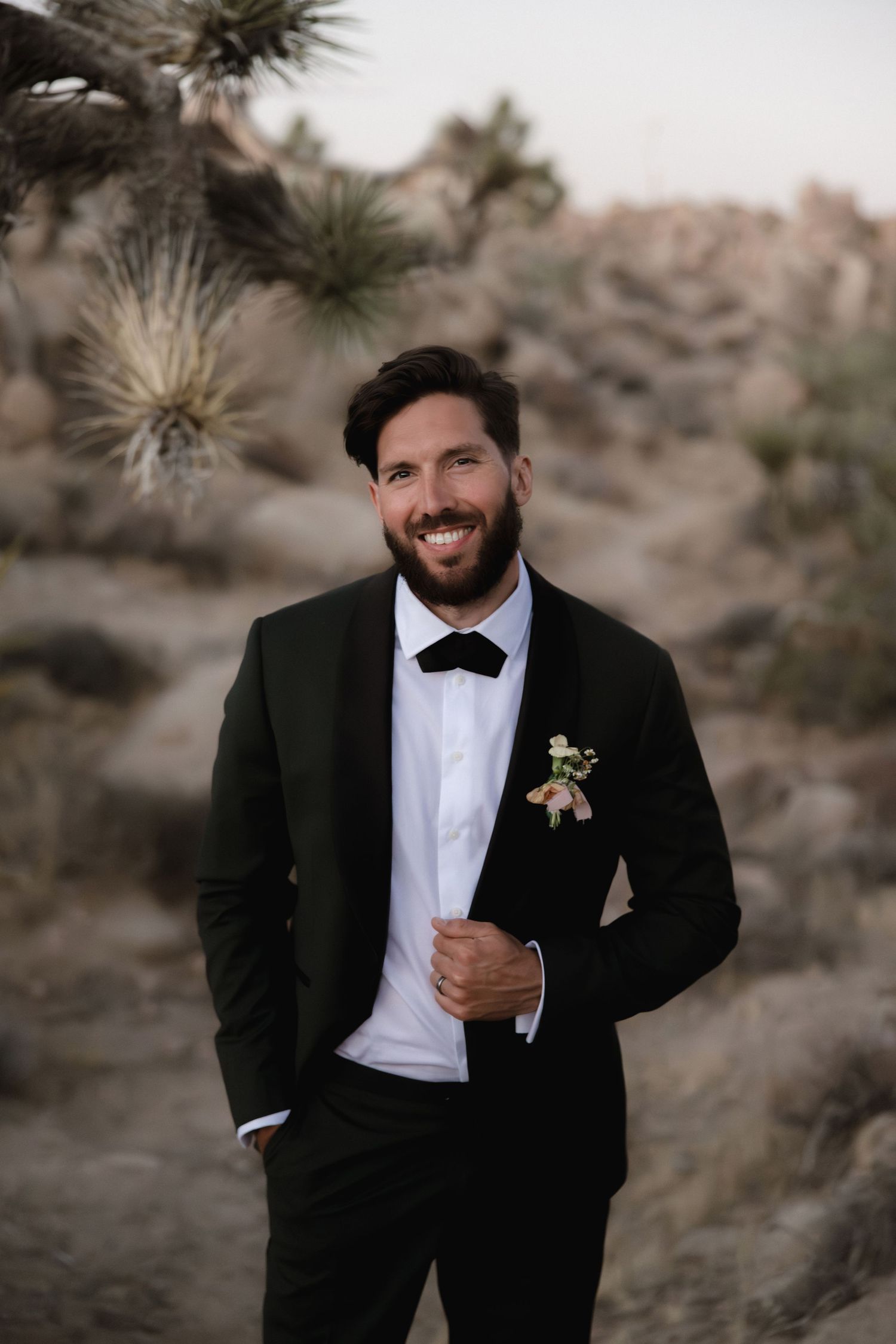 Groom in classic black tuxedo and white bow tie poses outdoors in a desert landscape at sunset.