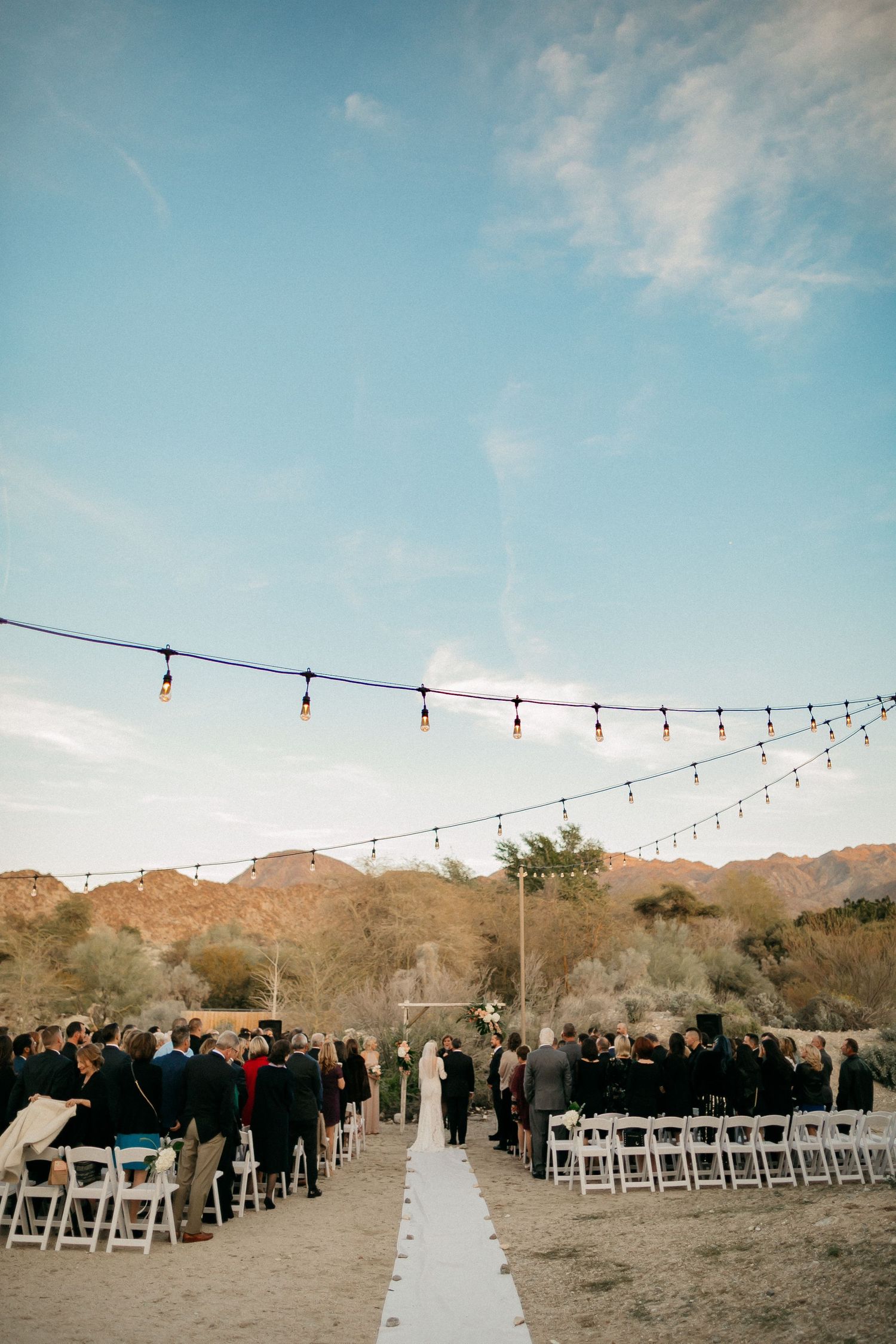 An outdoor desert wedding setup with white chairs and string lights under blue sky shows a ceremony path between seating.