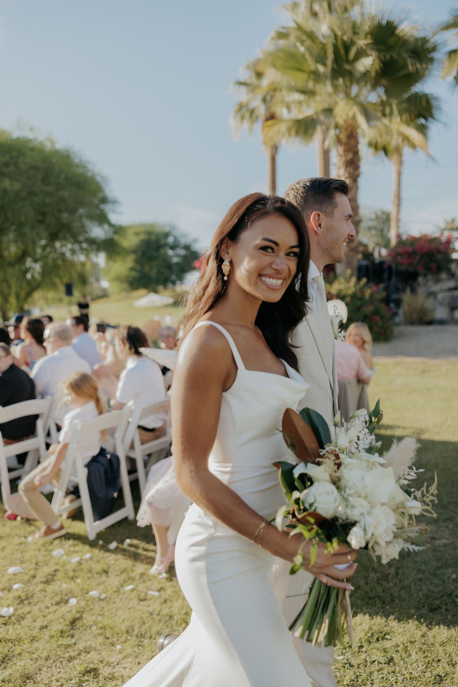 A joyful couple walks through a grassy outdoor florida wedding ceremony site surrounded by palm trees.