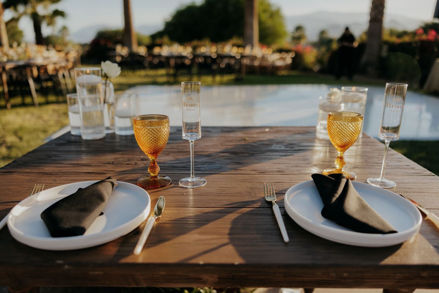 Place settings with wine glasses and white plates on rustic wooden table at sunset outdoor reception.