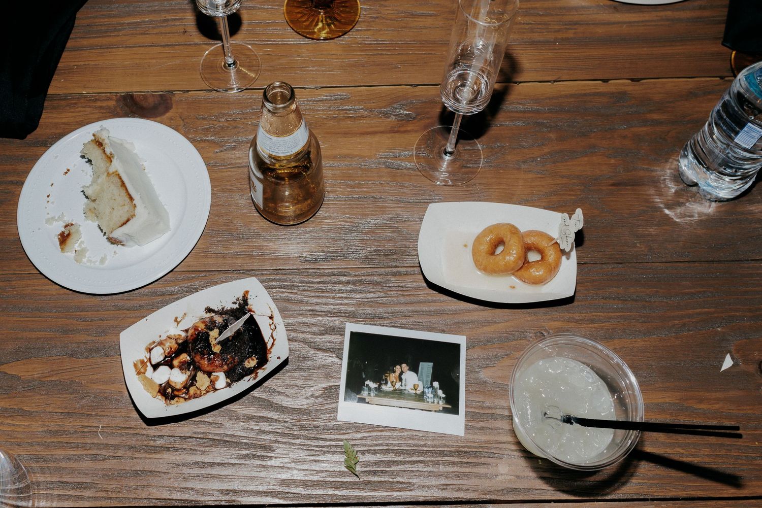 A wooden table with plates of food, drinks, cookies, and a polaroid photo laid out during a casual dining setting.