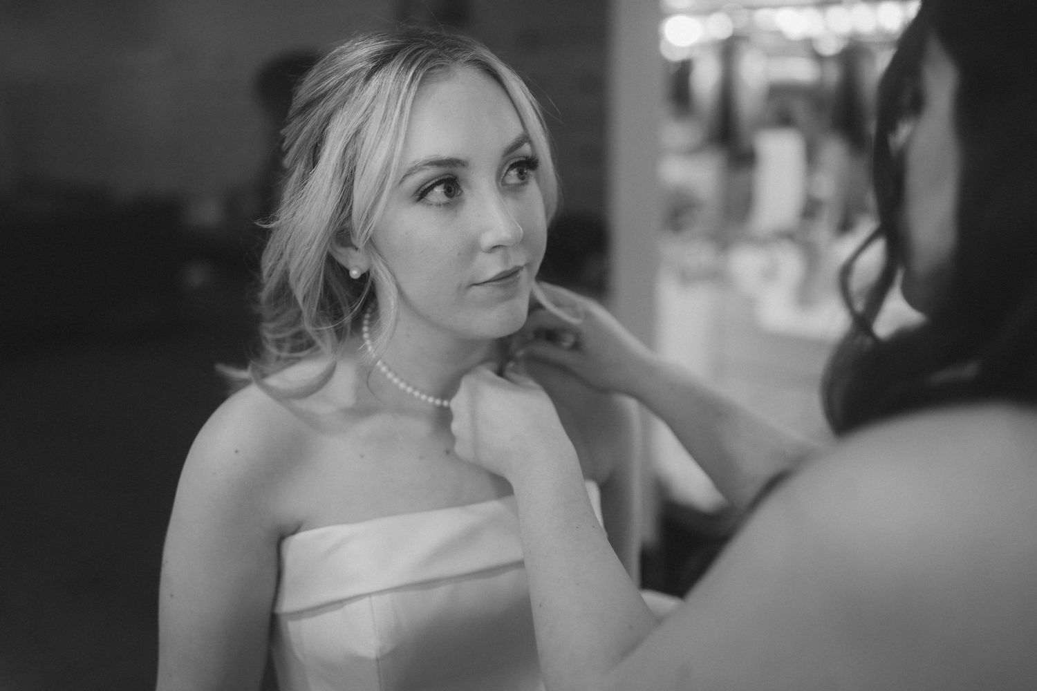 Black and white photo of a bride having her pearl necklace fastened by a bridesmaid before her wedding ceremony.