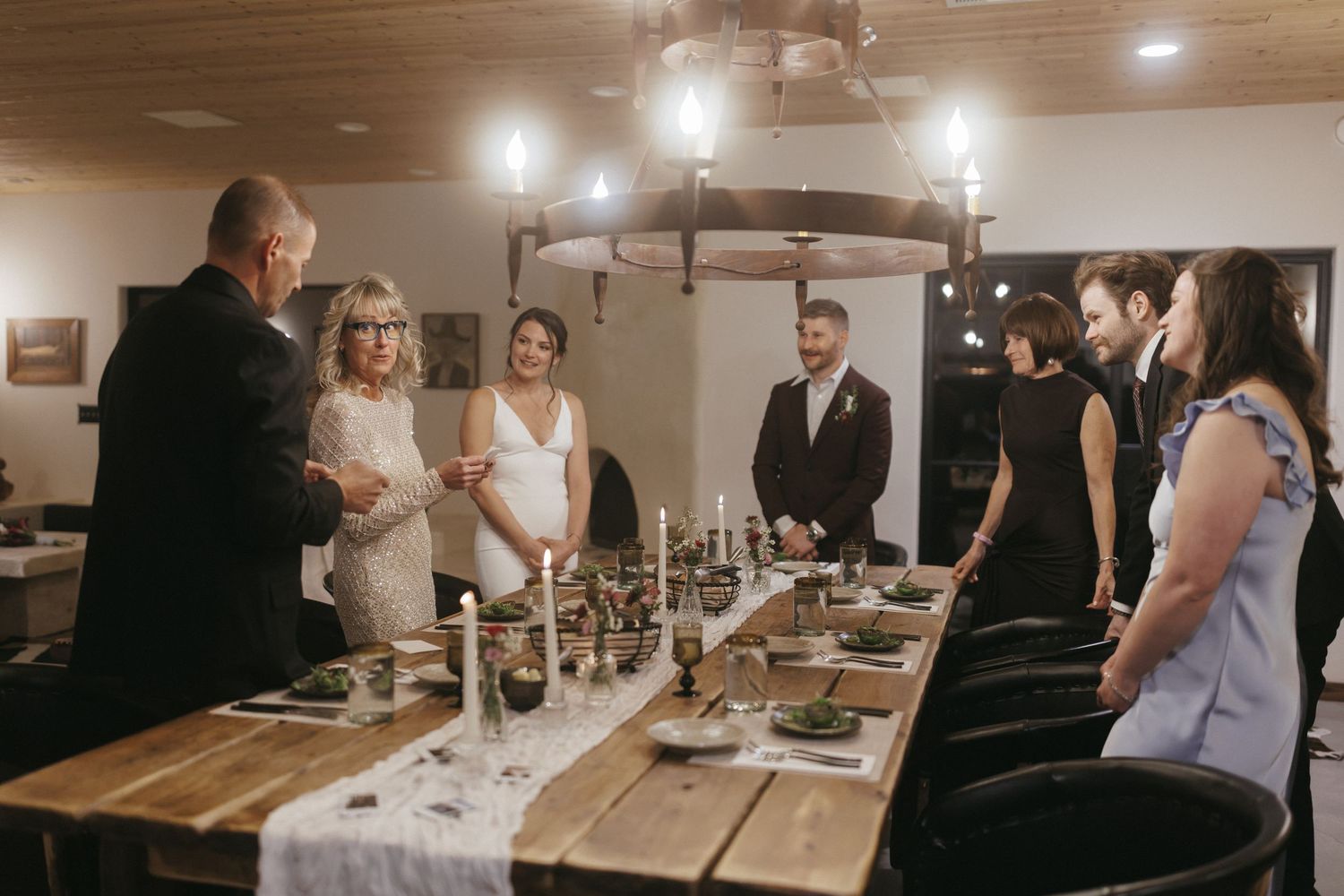 Wedding party gathers around rustic wooden dining table for intimate indoor reception celebration.