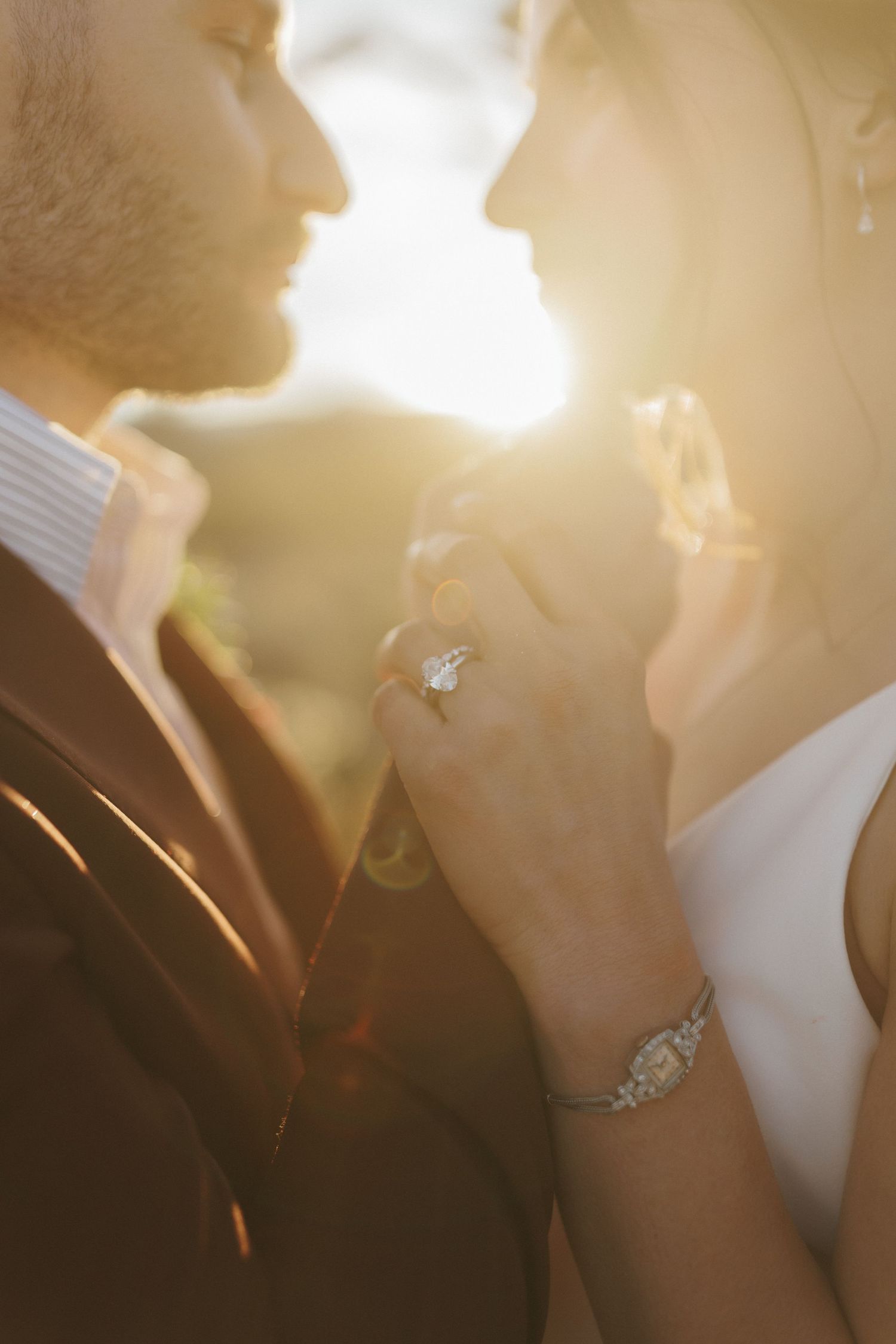 Bride and groom nearly kissing in golden sunset light, hands clasped showing diamond engagement ring.