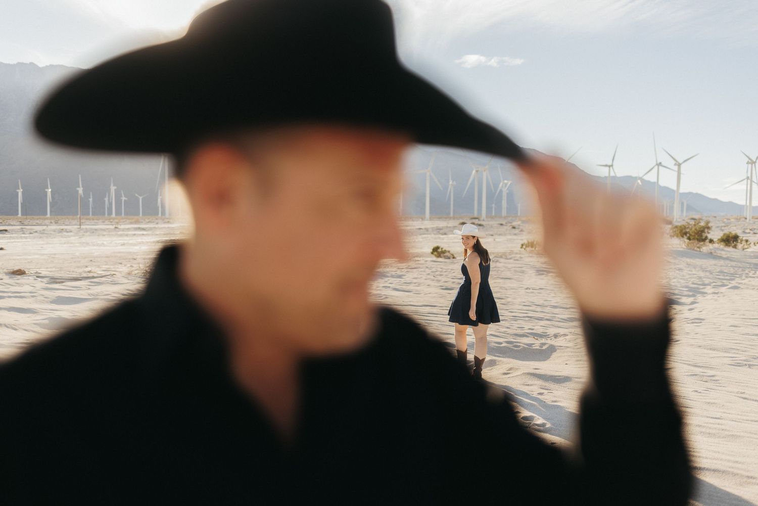 Man in black cowboy hat in desert with wind turbines, woman in black dress stands in background.