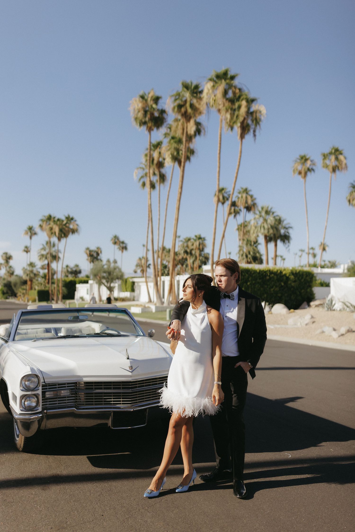 Couple in formal attire poses by a white vintage Cadillac convertible with palm trees in Palm Springs.