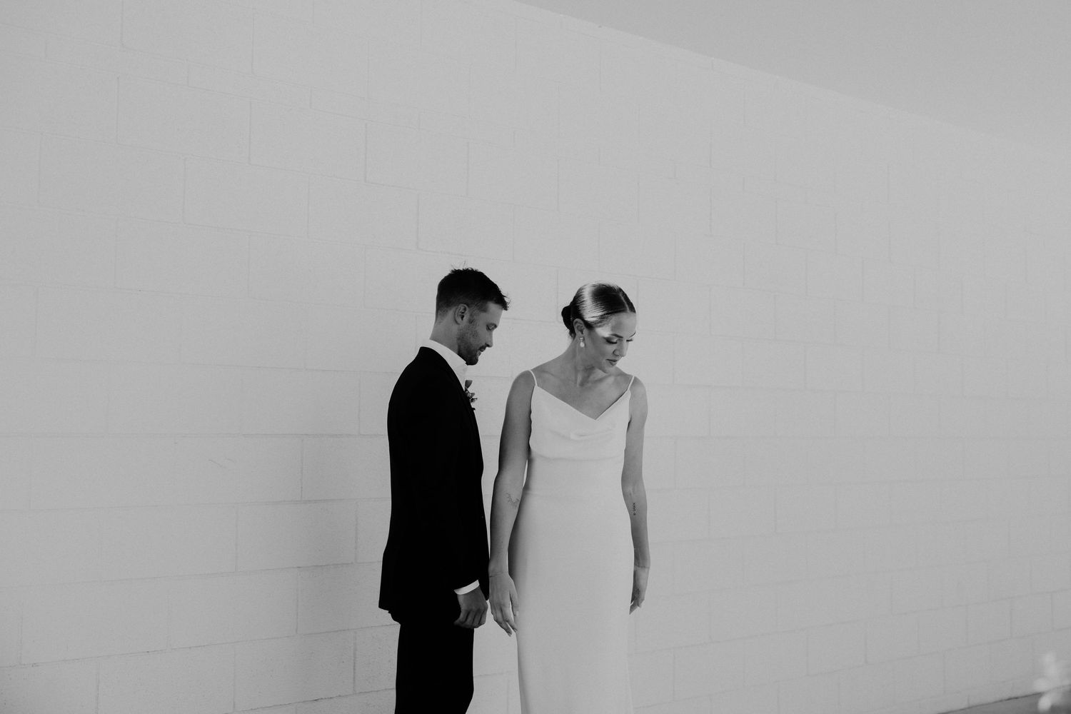 Black and white photo of a bride and groom holding hands, standing against a white wall in elegant wedding attire.