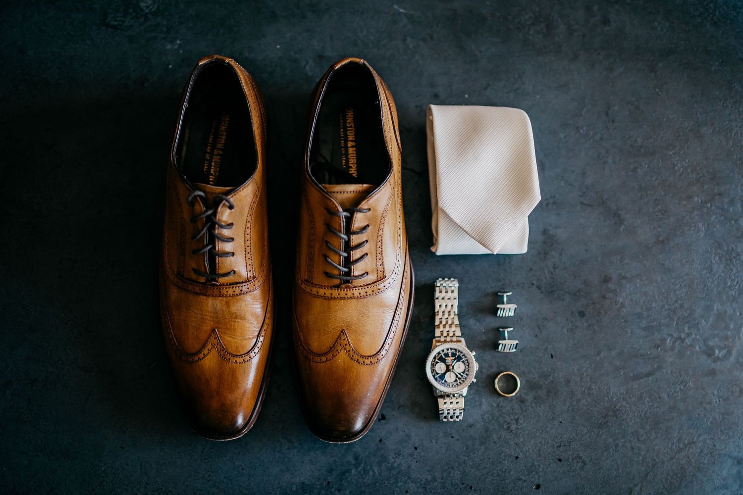 Brown leather dress shoes and watch accessories arranged against a dark gray backdrop for groom preparation.