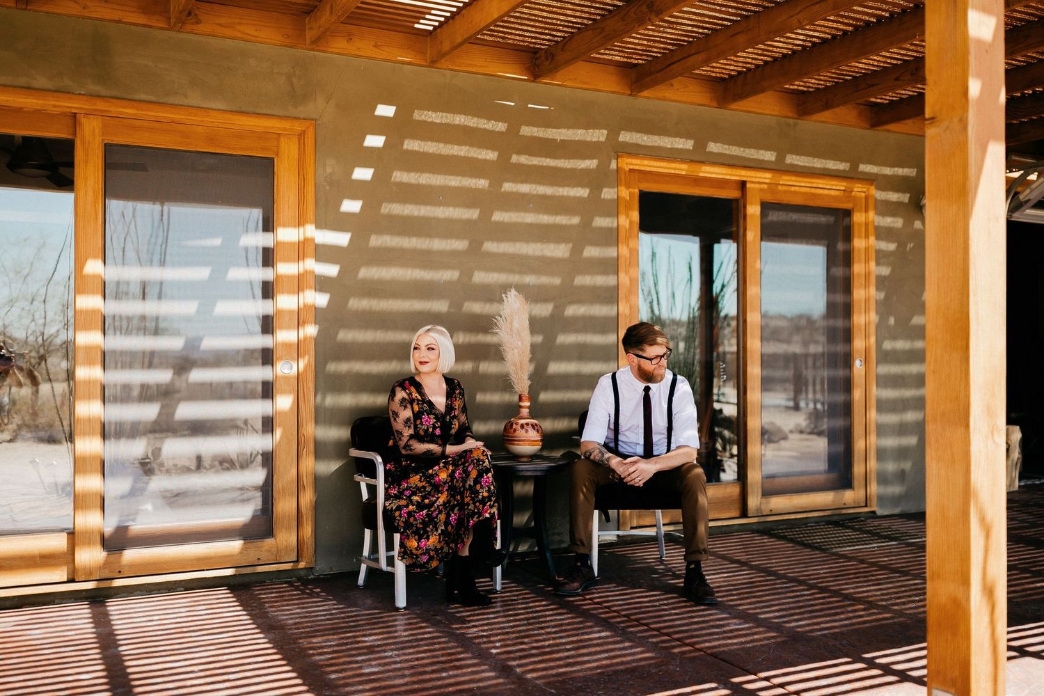 Two people enjoy a sunny afternoon on a rustic wooden porch with dramatic shadow patterns.